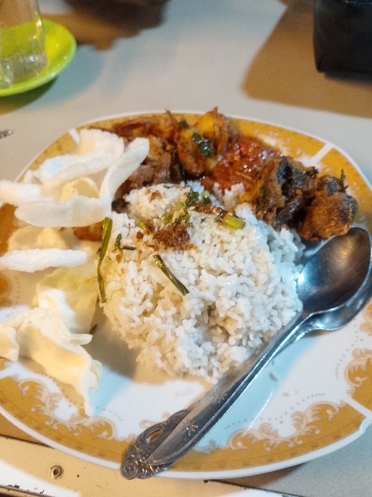 A close-up shot features a prepared meal served on a decorative plate, indicative of an indoor dining setting in Lhokseumawe, Indonesia. The main dish consists of a mound of white rice, lightly garnished with what appears to be fried shallots and chopped green herbs. To the right of the rice are several pieces of fried meat, possibly chicken, some coated in a reddish, glossy sauce, suggesting a savory and potentially spicy flavor. On the left side of the rice, several large, white, puffy crackers, known as krupuk, are arranged, a common accompaniment in Indonesian cuisine.
A silver-colored spoon rests on the right edge of the plate, extending over some of the food. The plate itself is white with a prominent gold or yellow rim, adorned with intricate, swirling ornamental patterns. The background is softly blurred, revealing a light-colored surface, likely a table, and in the upper left corner, a hint of a clear glass resting on a bright green saucer, suggesting a drink is also part of the meal. There are no individuals visible in the frame, nor is any text present. The scene captures a ready-to-eat meal, characteristic of local Indonesian food culture.