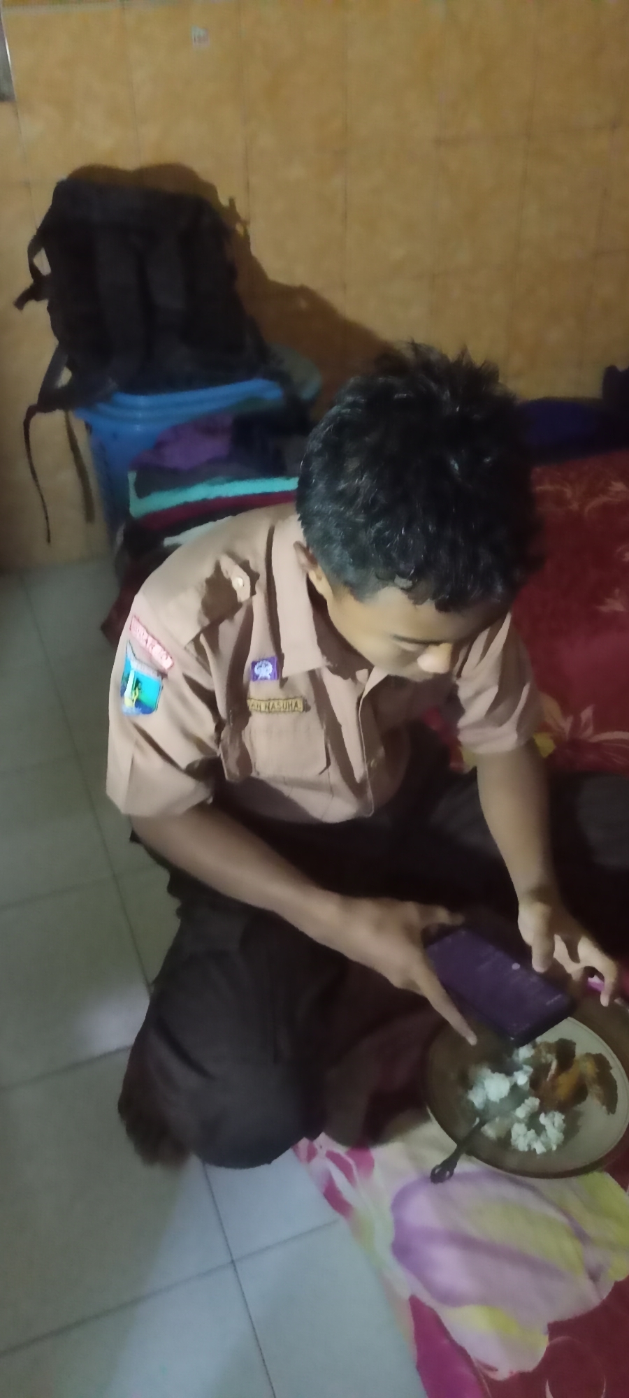 The image shows a young person, dressed in a brown uniform with patches on the chest, kneeling on a tiled floor in what appears to be a room in Gempolklutuk, Indonesia. The person is holding a dark-colored smartphone in their right hand and looking down at it. In front of the person, on a colorful blanket, is a plate of food, consisting of white rice and some pieces of dark meat, with a spoon resting in the plate. To the left of the person, there is a blue plastic bin filled with folded clothes, and behind that, a dark backpack is placed on a surface. The walls of the room are a light brown color with a tiled pattern. The lighting in the room is dim, suggesting it might be evening or nighttime. The overall scene depicts a casual moment of a person interacting with their phone while having a meal.