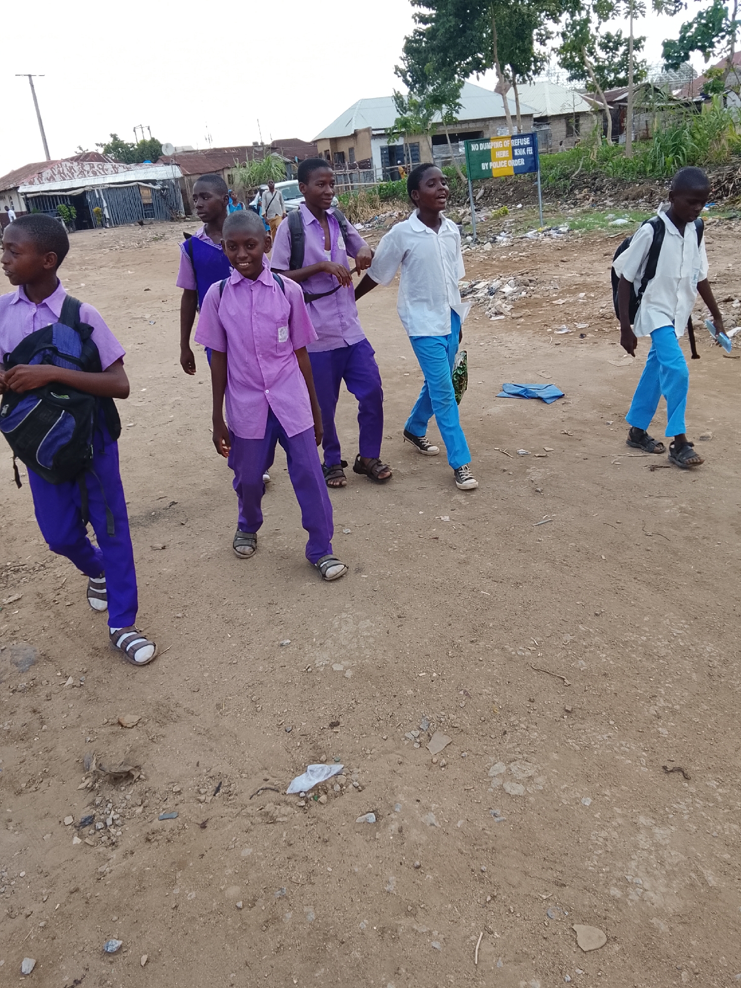 A group of six schoolboys, wearing school uniforms and backpacks, are walking on a dirt road. Four of the boys are wearing purple shirts and trousers, while two wear white shirts and blue trousers. The boys are in an environment with various residential buildings and a sign which reads: "NO DUMPING OF REFUSE HERE N30K FEE BY POLICE ORDER". The location is in Abuja, Nigeria.