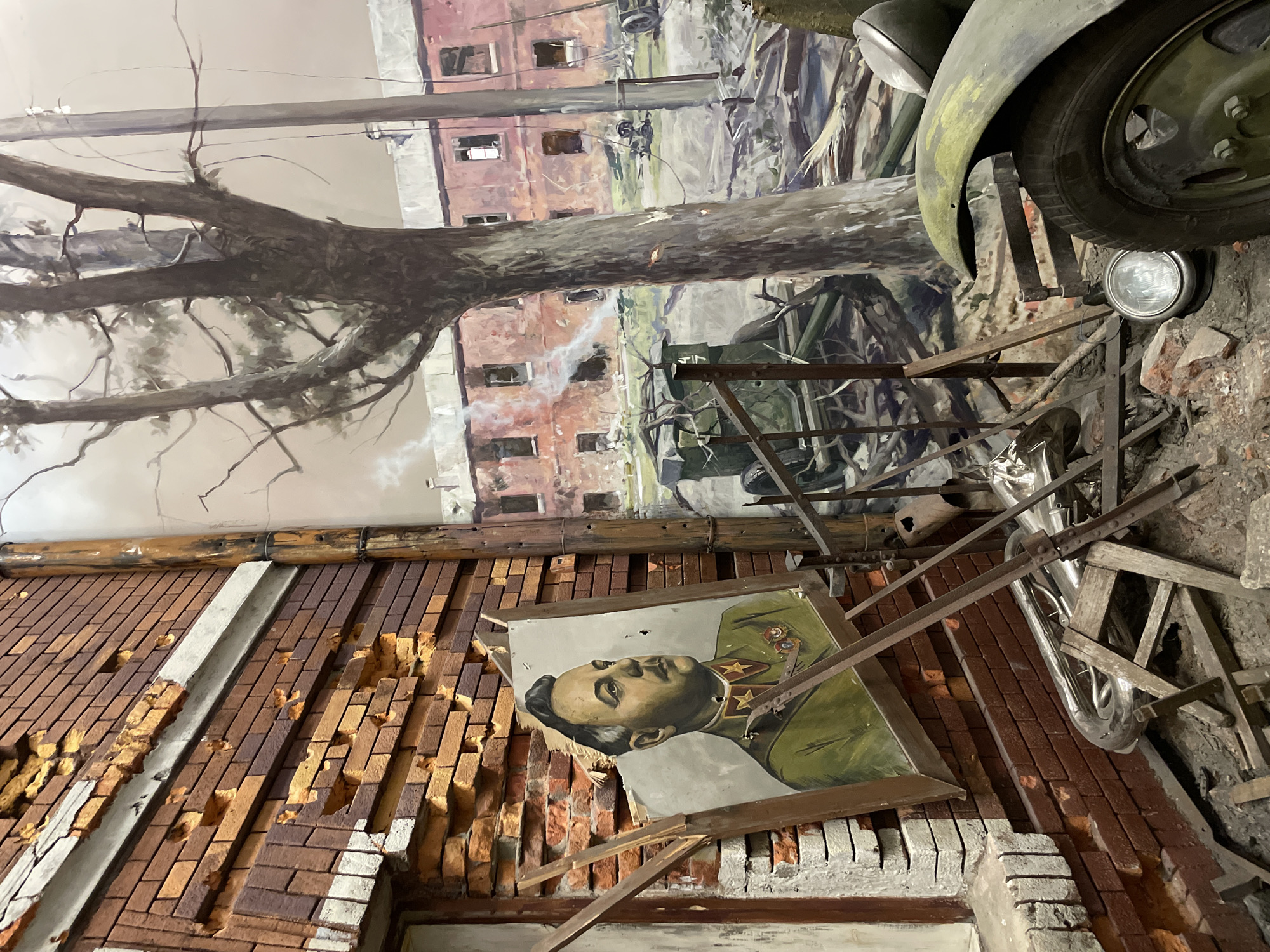 The image depicts a diorama exhibit likely portraying a scene of wartime destruction. A damaged brick wall with missing sections and mortar is prominent, with a portrait of a Soviet military officer resting against it, its frame broken. Above the wall, a mural of a destroyed urban environment is visible, showing damaged buildings and what appears to be a bare tree branch. A portion of a military vehicle, including a tire, fender, and headlight, is positioned in the foreground. Miscellaneous debris, including metal bars and what looks like a tuba, is scattered around the scene, contributing to the impression of ruin and disorder. The location in the city of Moscow, Russia is suggested by the content of the portrait and the overall theme of the diorama.