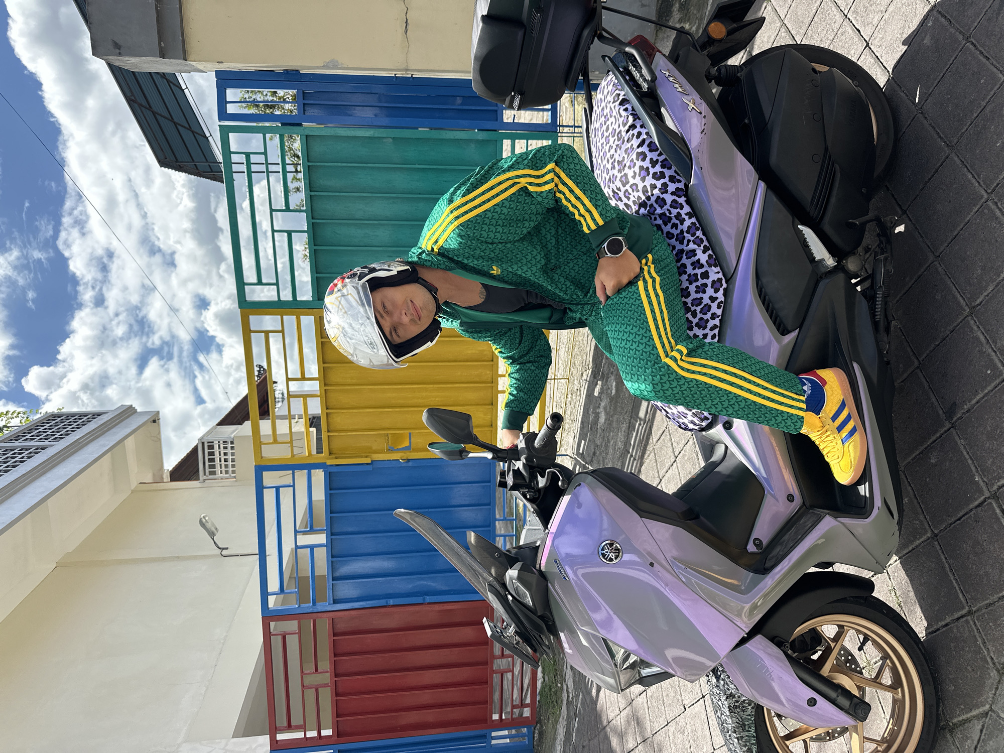 A medium shot depicts a person on a Yamaha scooter in North Kuta, Indonesia. The person, wearing a white helmet, green Adidas track suit with yellow stripes, and yellow Adidas sneakers with blue stripes, is positioned on the scooter, leaning slightly forward towards the handlebars. The scooter is silver-purple, with gold wheels and a black storage box attached behind the seat. The seat has a black and white spotted cover. Behind the person and scooter is a building with a stack of colorful rectangular metal frames in red, blue, yellow, and green. The sky is blue with scattered clouds. The ground is paved with gray bricks.
