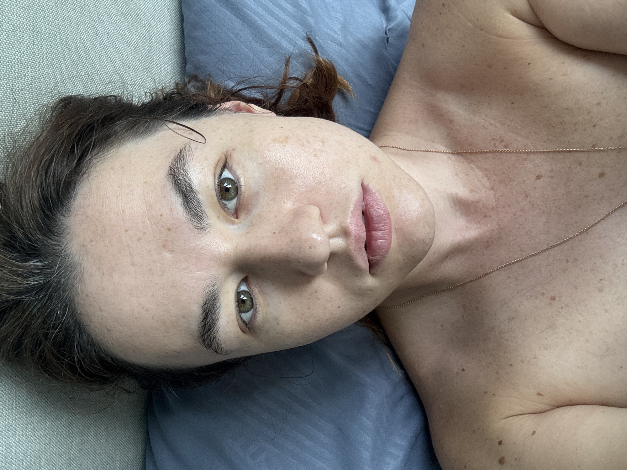 A young woman with brown hair, light skin, and green eyes is lying on a bed. She is wearing a gold chain necklace. The room appears to be a bedroom with a grey pillow and a textured wall. The image was taken in Ratsada, Thailand.
