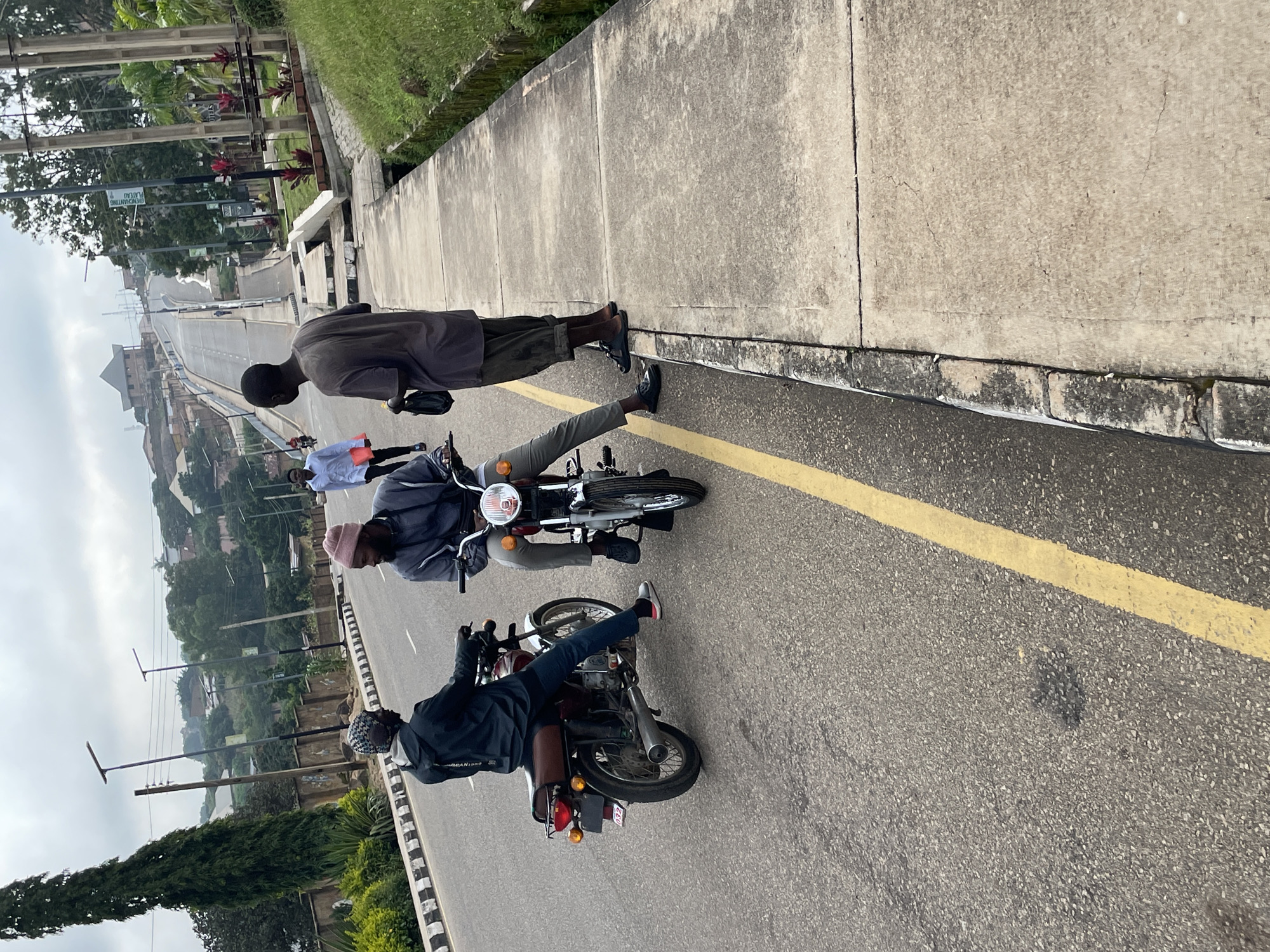 The image depicts a scene on a street in Jos, Nigeria. Two men are on motorcycles, facing each other on the road. One man, wearing a dark jacket and a patterned head covering, is seated on a red motorcycle. The other man, in a grey jacket and a pink beanie, is seated on a maroon motorcycle. To the right of the motorcycles, another man stands on the sidewalk, wearing a grey t-shirt and dark pants, holding a black bag. He is facing towards the motorcycles. In the background, the road slopes upwards, with buildings and vegetation visible on either side. Streetlights line the road. The weather appears overcast.