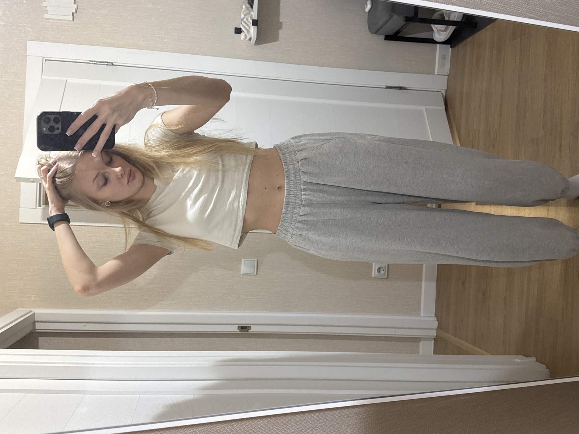 A young woman with blonde hair is taking a selfie in a mirror. She is wearing a white crop top and grey sweatpants. She has her hair pulled up and is looking down at her phone. The background appears to be an interior space, possibly a hallway or bedroom, with a door and a light-colored wall. There is a floor with wooden planks. The scene is consistent with an indoor environment in a residential setting. Without further information or visual cues, it is impossible to determine the specific location within the city of Perm, Russia.