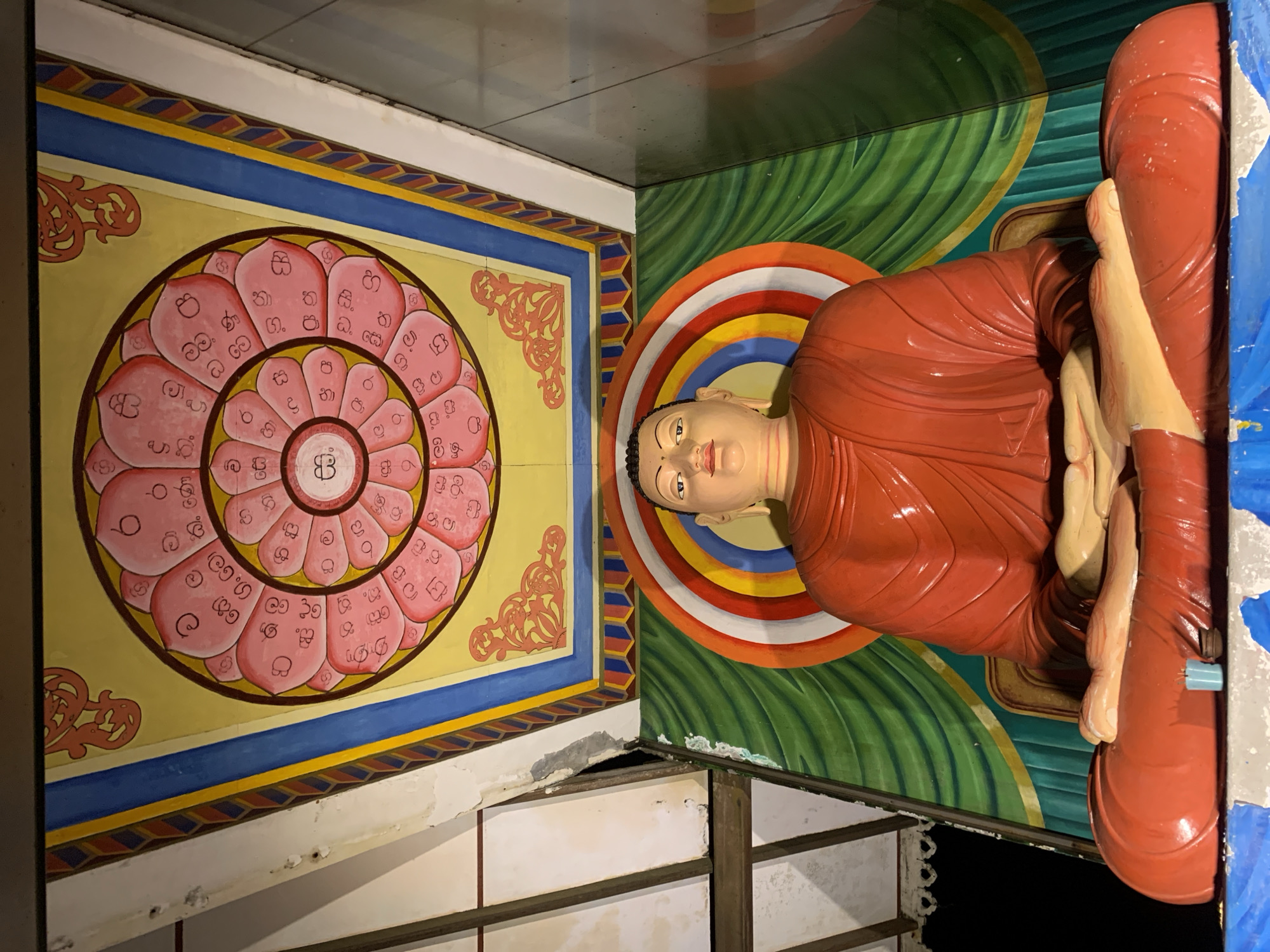 The image, taken in Kathaluwa, Sri Lanka, captures a vibrant and detailed section of a temple interior, featuring a reclining Buddha statue alongside ornate wall paintings. The Buddha is depicted lying on its right side, with its head gently resting on its right hand, embodying a state of serene meditation or Nirvana. The figure is clad in flowing reddish-orange robes, and its skin is a pale tone. A radiant, multi-colored halo, composed of concentric rings in red, orange, yellow, white, and blue, frames the back of the Buddha's head, set against a background of rich green, undulating patterns that suggest foliage.
Adjacent to the Buddha, a large, decorative mural dominates the wall. It showcases a magnificent circular motif, resembling a stylized pink lotus flower or mandala, painted on a vivid yellow ground. Within the petals of this lotus, numerous symbols and characters are visible, primarily appearing as Sinhala script, consistent with the regional context. This mural is bordered by a distinct blue band and an intricate patterned strip featuring geometric designs in varying hues. Below the elaborately painted sections, parts of the building's structure show signs of age or wear, with patches of exposed material and visible framework, contrasting with the vibrant artwork above. The overall scene conveys a strong sense of cultural and religious artistry.