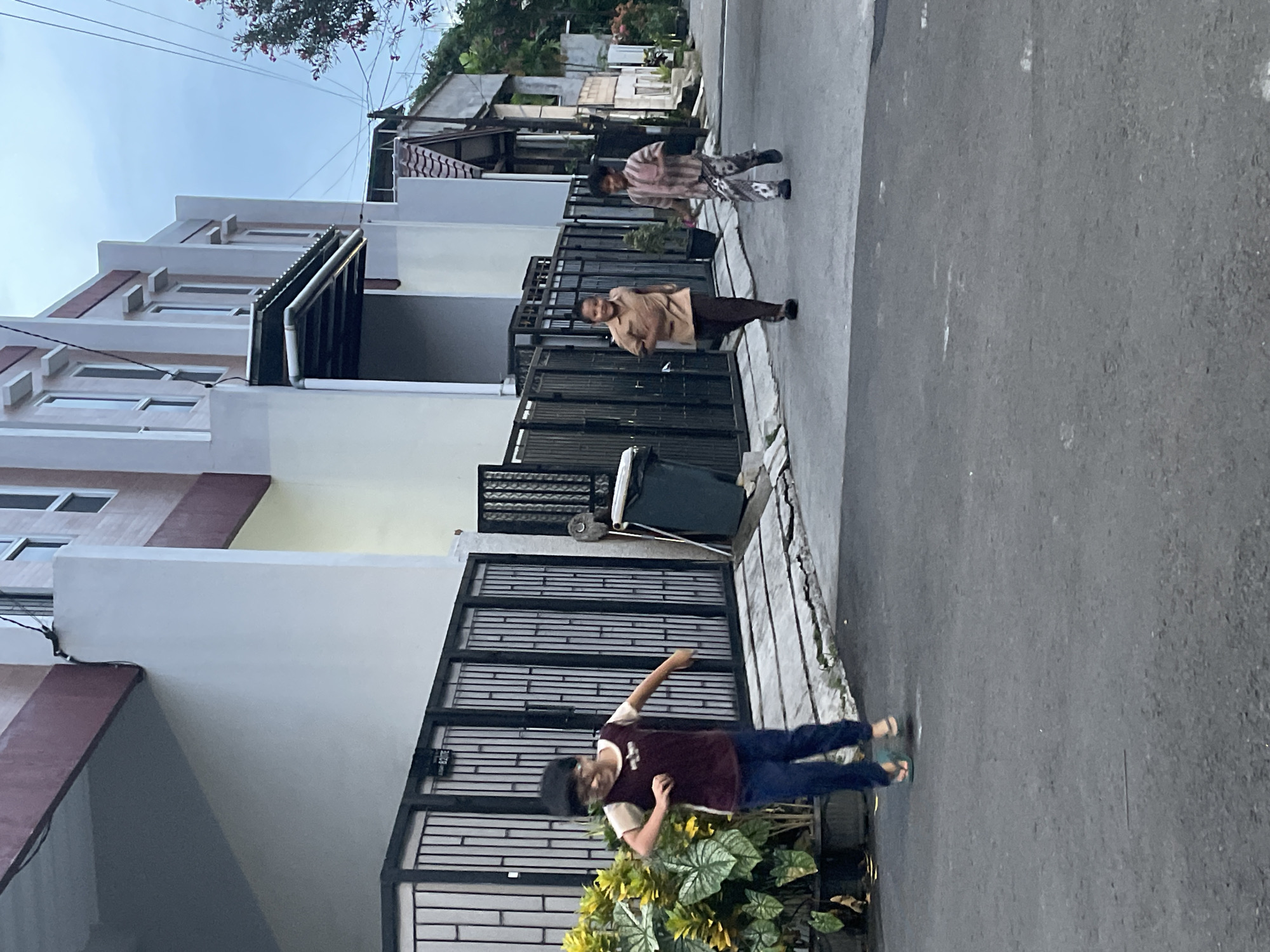 The image depicts a street scene in a residential area. The location is North Jakarta, Indonesia, evidenced by the architecture of the houses and the presence of local residents. Three individuals are visible: a child in the foreground, wearing a dark shirt and jeans, gesturing with their arm; an adult wearing a tan shirt and dark pants; and another person wearing a striped shirt and patterned pants. They are walking along the sidewalk. The houses are two-story structures with gated frontages. A green garbage bin is located between two of the houses. The street is paved, and foliage is visible in front of one of the houses. The sky is overcast.
