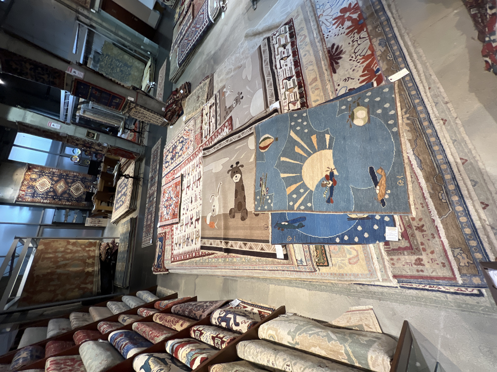 The image depicts the interior of a rug store, possibly in Yerevan, Armenia, based on the style of rugs displayed. The scene is oriented in a way that the walls are viewed horizontally. Rugs of various designs and sizes are hung on the walls. A display rack with rolled-up rugs is visible along the bottom edge of the image. No people are clearly visible in the main viewing area, but some dark figures are discernible near the door on the left. The store seems to be well-lit, likely from both natural and artificial sources.