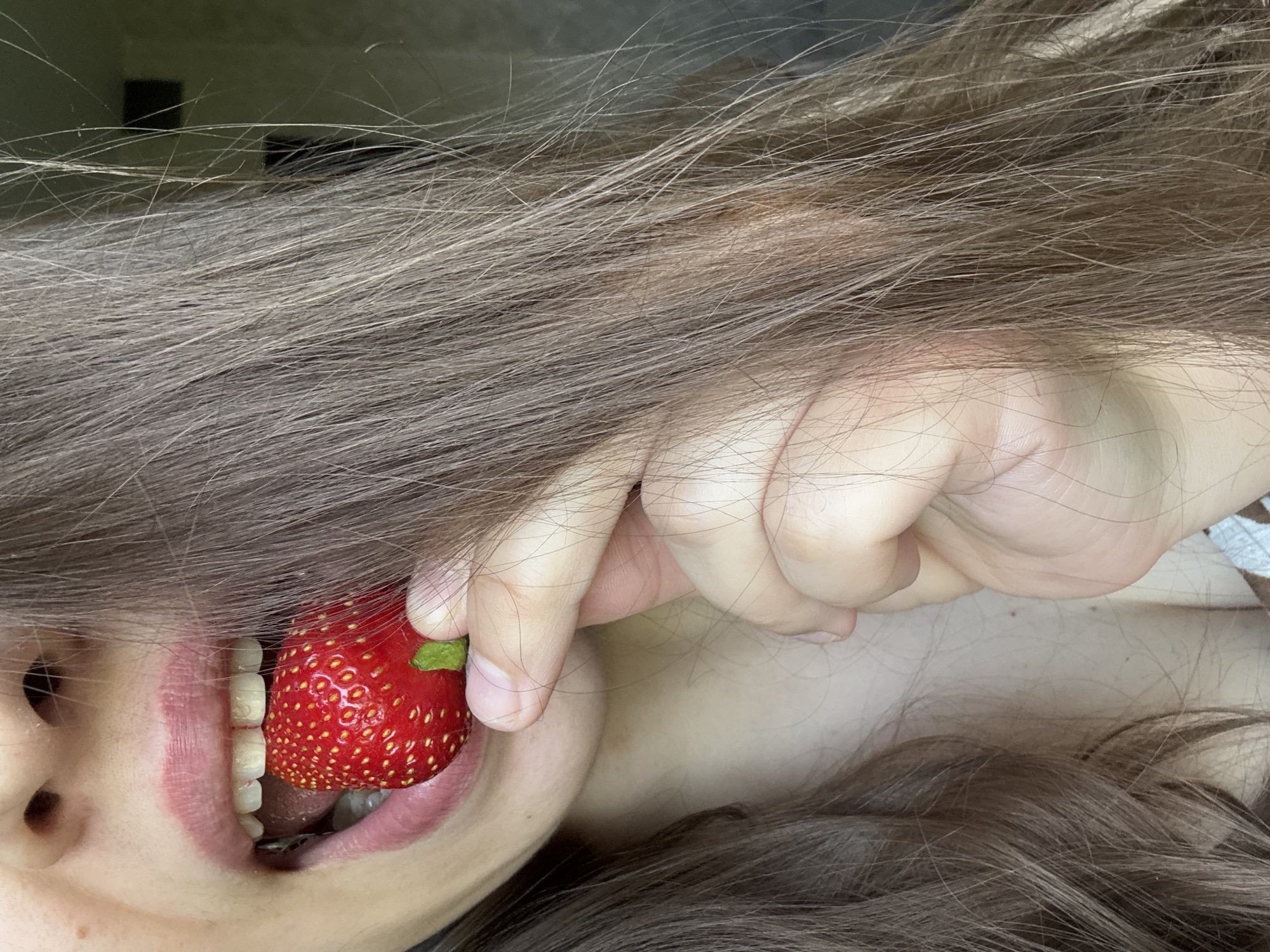 A close-up, outdoor, natural light portrait shows a person's face and upper chest. The person has long, brown hair and is holding a ripe strawberry to their mouth. Their mouth is open, and their teeth are visible as they bite into the fruit. The background is blurred, but it appears to be an indoor setting with a plain wall. The image does not contain any elements that would suggest a specific location in Krasnoyarsk, Russia.