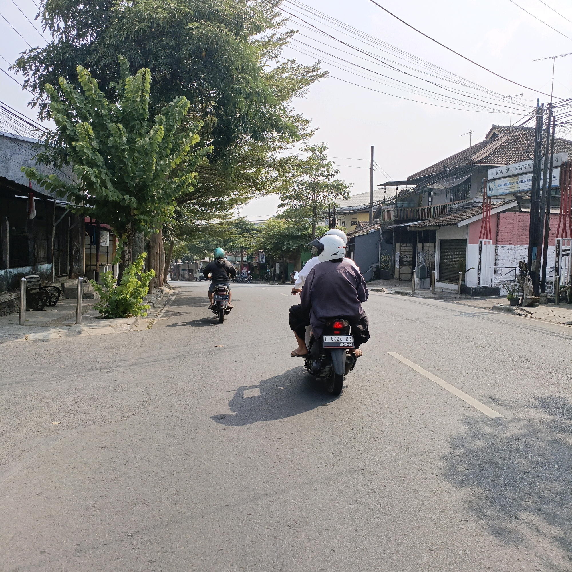 The image captures a street scene in Salatiga, Indonesia, on a bright, sunny day. The foreground features an asphalt road with a white dashed line dividing the lanes. In the middle ground, three motorcycles are visible. The closest motorcycle carries two individuals wearing helmets, with the rear passenger in a dark purple jacket and the driver in white. This motorcycle has a license plate that reads "H 6424 RR". Another motorcycle further ahead has a single rider wearing a green helmet, with the license plate "H 3721 TC". A third motorcycle is parked on the side of the road in the distance.
The environment includes buildings lining the street, some with tiled roofs and open storefronts. Several trees with lush green foliage are scattered along the roadside, providing shade. Utility poles with numerous wires stretch across the sky, indicating an urban or suburban setting. The overall impression is one of daily life on a moderately busy street in an Indonesian city.