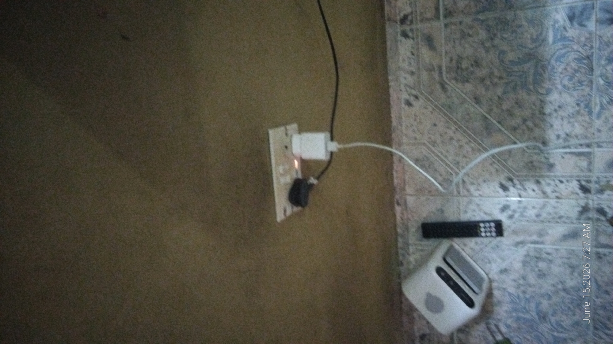 The image captures an indoor scene with a focus on electrical outlets and devices on a textured wall and a tiled floor. In the upper portion of the image, a white electrical outlet strip is mounted on a beige wall. A black electrical adapter is plugged into one socket, and a white power adapter for a device is plugged into another socket. A white cable extends from the white power adapter, and a black cable is also connected to the black adapter. The lower portion of the image shows a section of a tiled floor. On the floor, there is a white electronic device with a grill-like opening and an Apple logo, likely a speaker or router. Next to it, a black remote control with multiple buttons is placed. The overall environment appears to be a domestic setting, likely a room in a house or apartment. There is a timestamp in the lower-left corner of the image that reads "June 15, 2026 7:27 AM". The location is identified as Howayi, Nigeria.