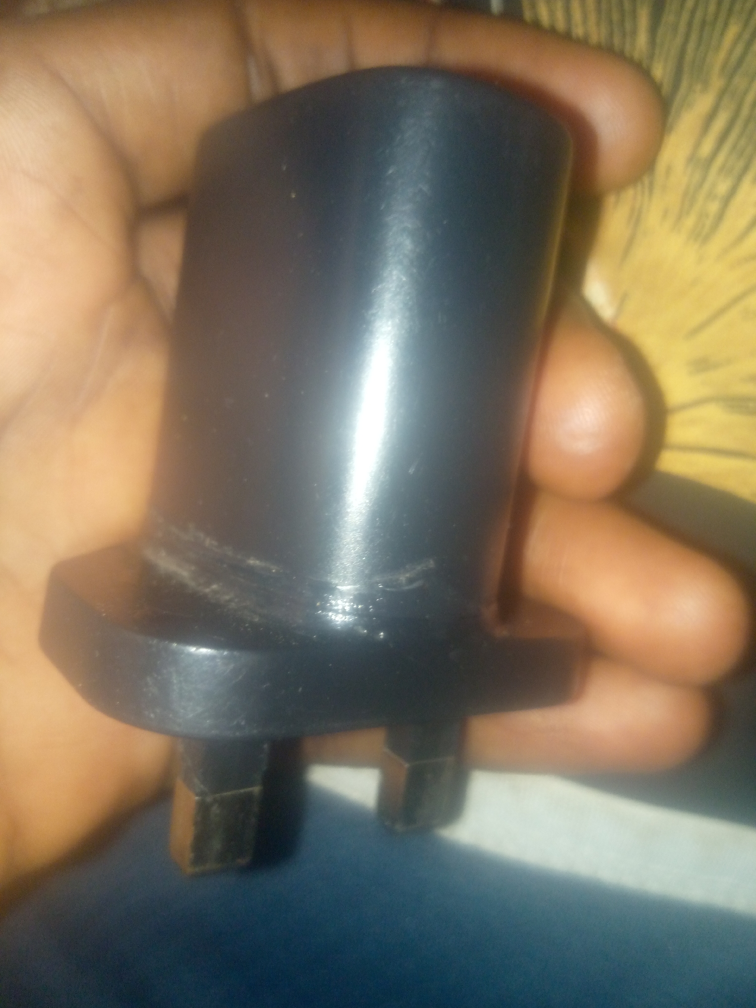 An extreme close-up, low-angle shot shows a hand holding a black electrical plug. The plug has a wide, flat base from which three rectangular pins extend downwards. The pins appear to be made of a metallic material with a copper-colored hue. The hand holding the plug is dark-skinned, and the thumb and index finger are clearly visible, gripping the base of the plug. The background is blurred, with a yellow, textured surface visible on the upper right, and a dark blue surface below. The lighting is uneven, creating highlights on the glossy surface of the plug. The image is grainy and lacks detail, making it difficult to ascertain the precise type of plug or its condition beyond its color and shape. There is no discernible context that would place this image in the city of Jauro Yinu, Nigeria.