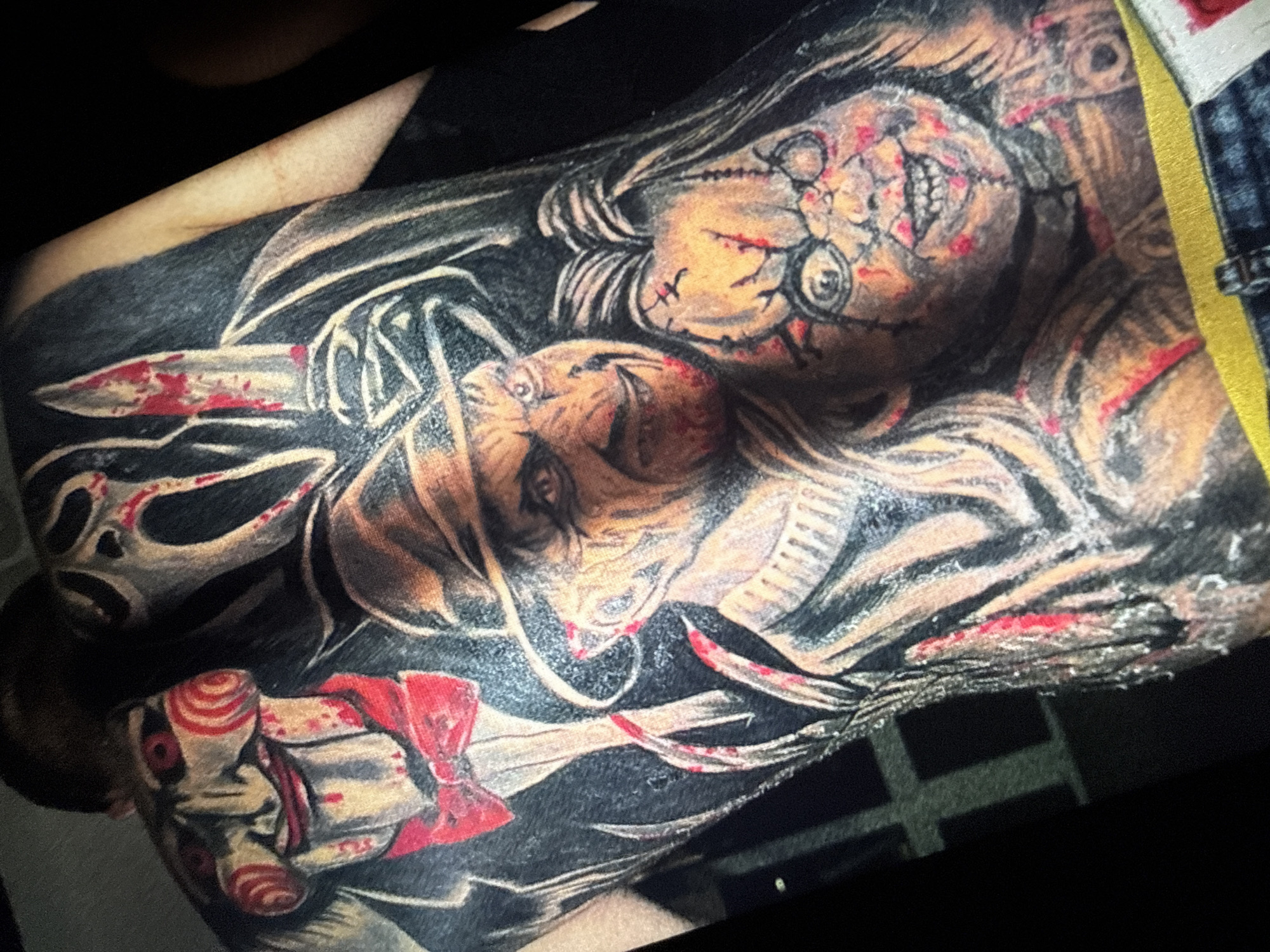 The image shows a detailed tattoo on a person's back, featuring several iconic horror movie characters. From left to right, the characters depicted are Billy the Puppet from the Saw franchise, Ghostface from the Scream franchise, Freddy Krueger from the Nightmare on Elm Street franchise, and Chucky from the Child's Play franchise. The tattoo is rendered in a realistic style with a focus on the characters' menacing expressions and signature elements, such as bloodstains and weapons. The person is wearing denim jeans at the bottom of the frame. The background is dark and out of focus, suggesting the image was taken indoors. There is no discernible information in the image that would indicate its location in Sofia, Bulgaria, or any other specific city.