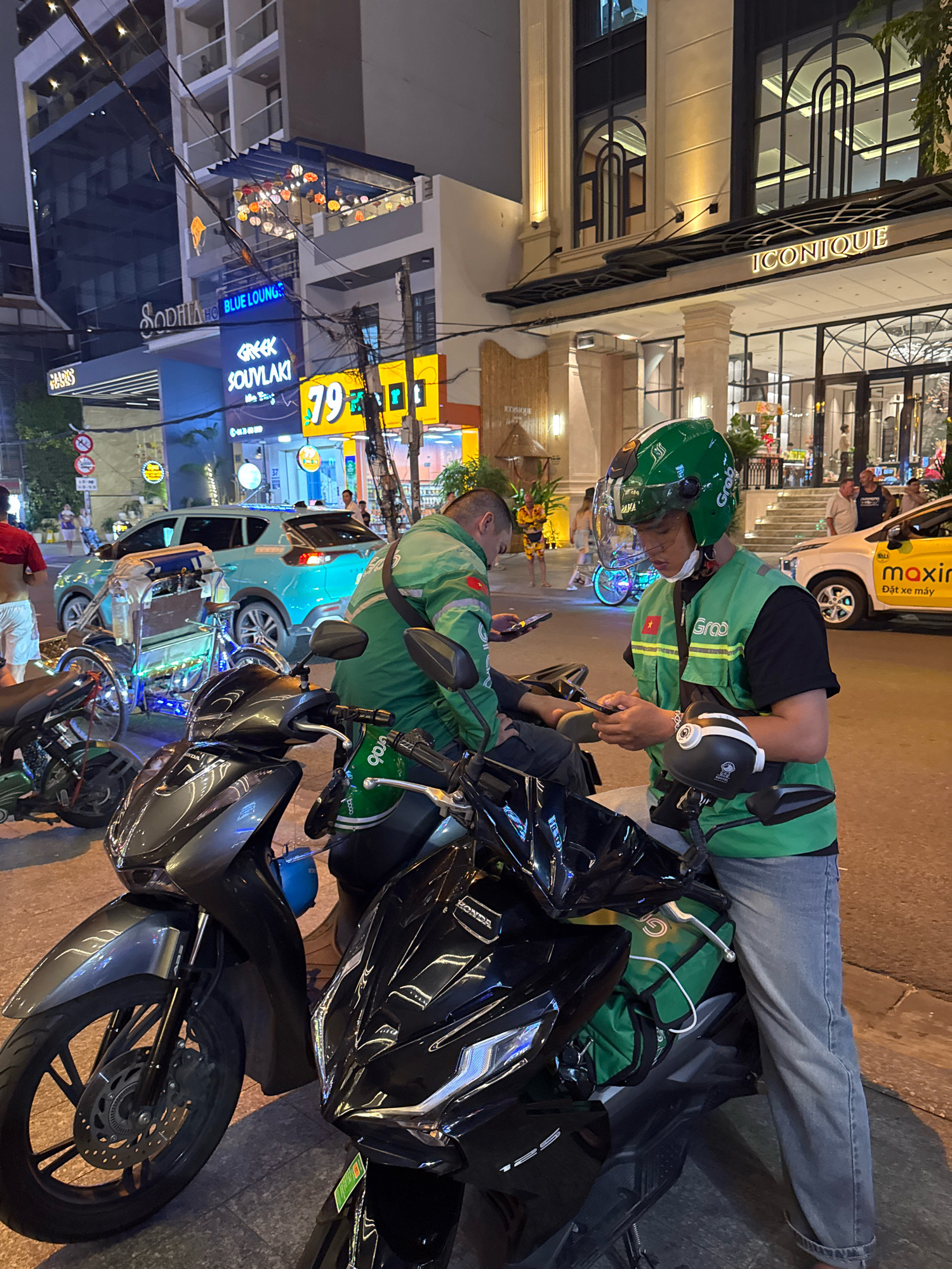 A street scene at night in Nha Trang, Vietnam, captures two Grab food delivery riders. The rider in the foreground, wearing a green helmet with a clear visor and a green vest over a black t-shirt, is looking down at his phone while holding it with both hands. He is seated on a black motorcycle, with another dark grey motorcycle to his left. His left leg is visible, clad in blue jeans. A green Grab delivery bag is strapped to the back of the black motorcycle. To the left of the foreground rider, a second Grab rider, also wearing a green vest, is partially visible, looking at his phone. The background shows a street with buildings, including one with a sign for "Sophia Hotel BLUE LOUNGE" and another with "ICONIQUE" above its entrance. A turquoise car is parked on the street, and a decorated three-wheeled vehicle with blue lights is also present. Streetlights and neon signs illuminate the scene.