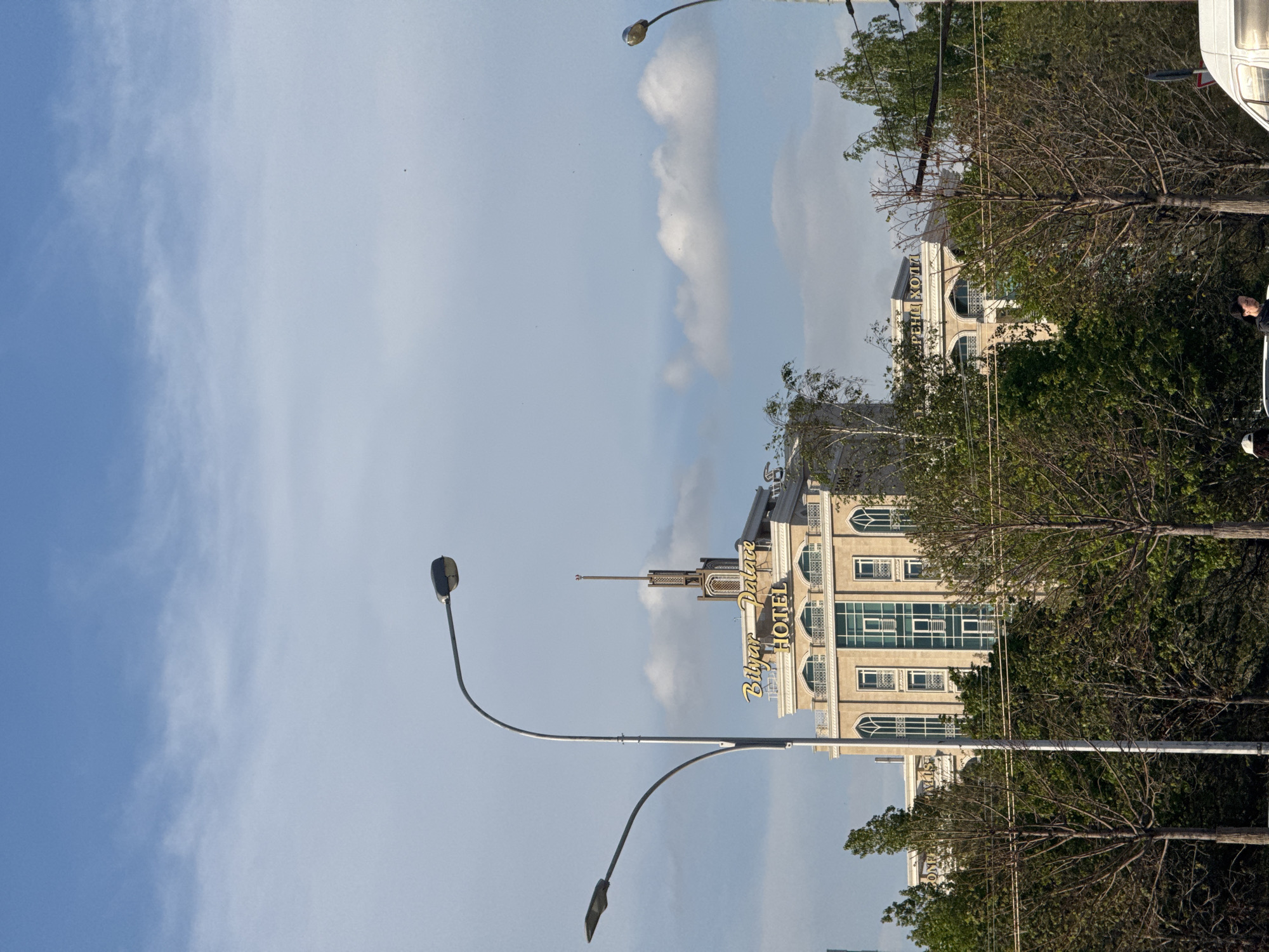 The image shows the Bilyar Palace Hotel in Kazan, Russia. The hotel is a tall, multi-story building with light-colored facade and many windows. There are trees and bushes in front of the hotel, partly obscuring the view. A street lamp is in the foreground. The sky is blue with some clouds. A portion of another building can be seen obscured by a tree. A vehicle is partially visible at the top right.