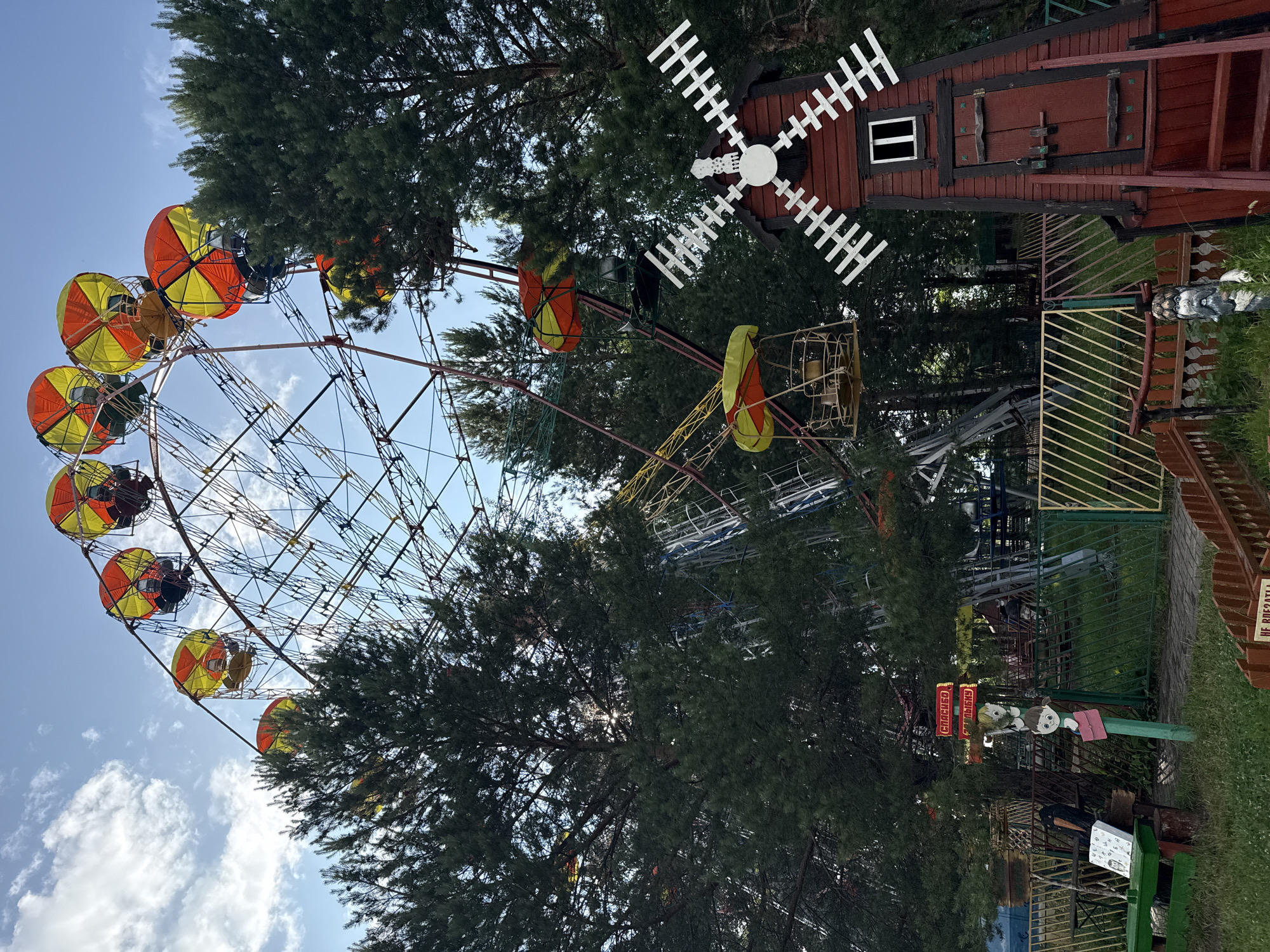 In an outdoor amusement park, a large Ferris wheel with yellow and orange gondolas is set against a cloudy blue sky and surrounded by tall green pine trees. To the right of the Ferris wheel, a small, red, wooden windmill stands with white blades and a dark door. In the foreground, a green picnic table sits on the grass, and a fence with yellow and green bars encloses a pathway. A signpost with images of children and red signs is visible near the picnic table. The ground is covered in grass and gravel, with a wooden fence bordering the right side of the image.