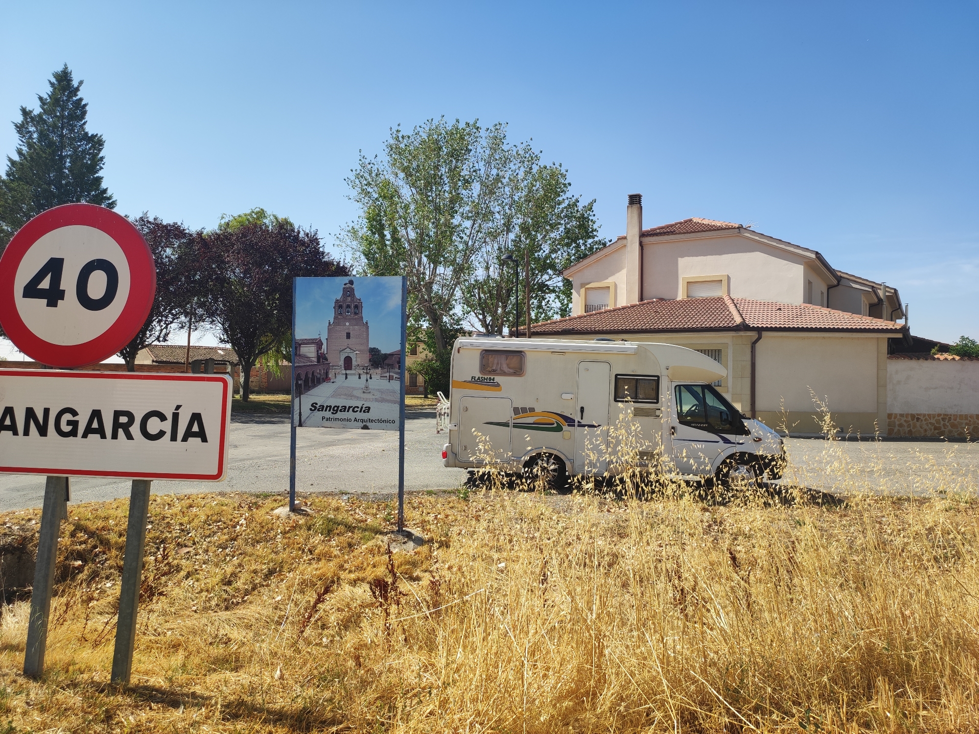 The image captures a roadside scene in Sangarcía, Spain, on a clear, sunny day. In the foreground, a white sign with a red border displays the word "SANGARCÍA" in black capital letters. To the left of this sign is a circular speed limit sign with a red border and the number "40" in black, indicating a speed limit of 40 kilometers per hour. Behind these signs, a residential building with a tiled roof is visible. To the right, a white recreational vehicle (RV) is parked on a paved area. The ground is covered with dry, golden grass and some scattered leaves. In the background, there are trees, including a tall evergreen tree and a tree with dark red foliage, as well as a building with a bell tower, suggesting a church or a historical structure. The overall environment appears to be rural or semi-rural.