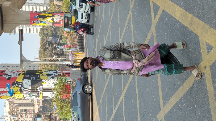 A man in worn clothes, including a tattered jacket and sandals, stands on a street marked with yellow intersecting lines. He wears a purple shirt and a lungi-style skirt. The background features a cityscape with buildings, street art, and a bridge structure. Cars and a motorcycle are visible on the street behind him.