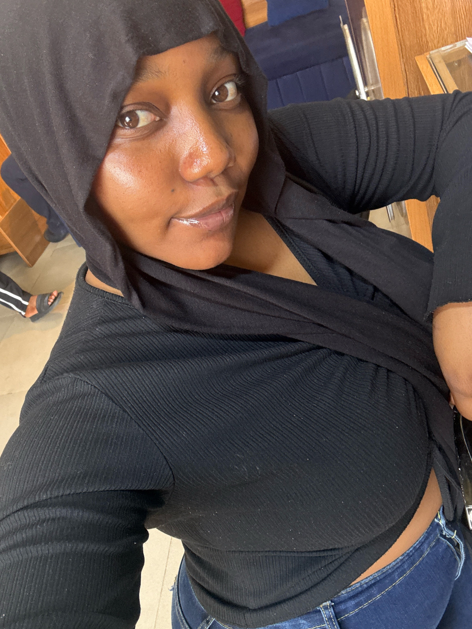 A high-angle medium shot captures a Black woman wearing a black hooded garment and a black ribbed crop top, paired with blue jeans. She is positioned in the foreground, facing slightly to her right and looking directly at the camera. Her hair is covered by the black hood, and she has on lip gloss, with subtle sheen on her cheekbones. In the blurred background, a portion of a navy blue couch and wooden furniture can be seen, suggesting an indoor setting. To the left of the frame, a person's legs are visible, one wearing a black sandal and striped pants, indicating the presence of others in the vicinity. The lighting is bright, illuminating the woman's face and highlighting the texture of her clothing. The scene does not provide specific visual cues to pinpoint its location within Jos, Nigeria.