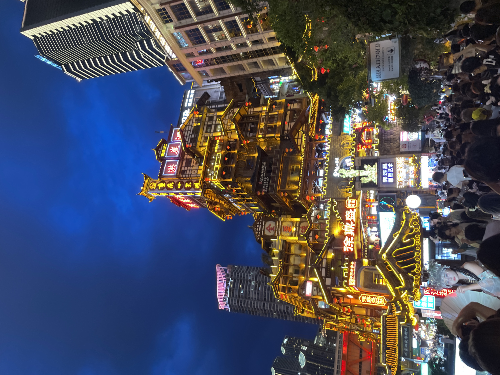 The image depicts a bustling night scene in the Yuzhong District of China, likely a vibrant commercial or tourist area. The focal point is a multi-tiered, traditional-style building adorned with numerous golden yellow lights and red lanterns, creating a warm, festive atmosphere. This structure is surrounded by modern high-rise buildings, some with illuminated facades and windows.
In the foreground, a dense crowd of people is visible, many holding up their phones, suggesting they are taking pictures or videos. In the center of the crowd, a young woman dressed in a white and blue outfit and an elaborate headdress stands out, holding a white parasol. She appears to be posing or interacting with the onlookers.
Various neon signs and illuminated billboards with Chinese characters are scattered throughout the scene, indicating commercial activity and advertising. A sign with English text, "ENTRANCE GATE 7," is also visible, along with arrows indicating directions and operating hours. The sky above is a deep blue, characteristic of twilight or nighttime. The overall impression is one of lively urban nightlife, a blend of traditional and modern architecture, and a significant gathering of people.