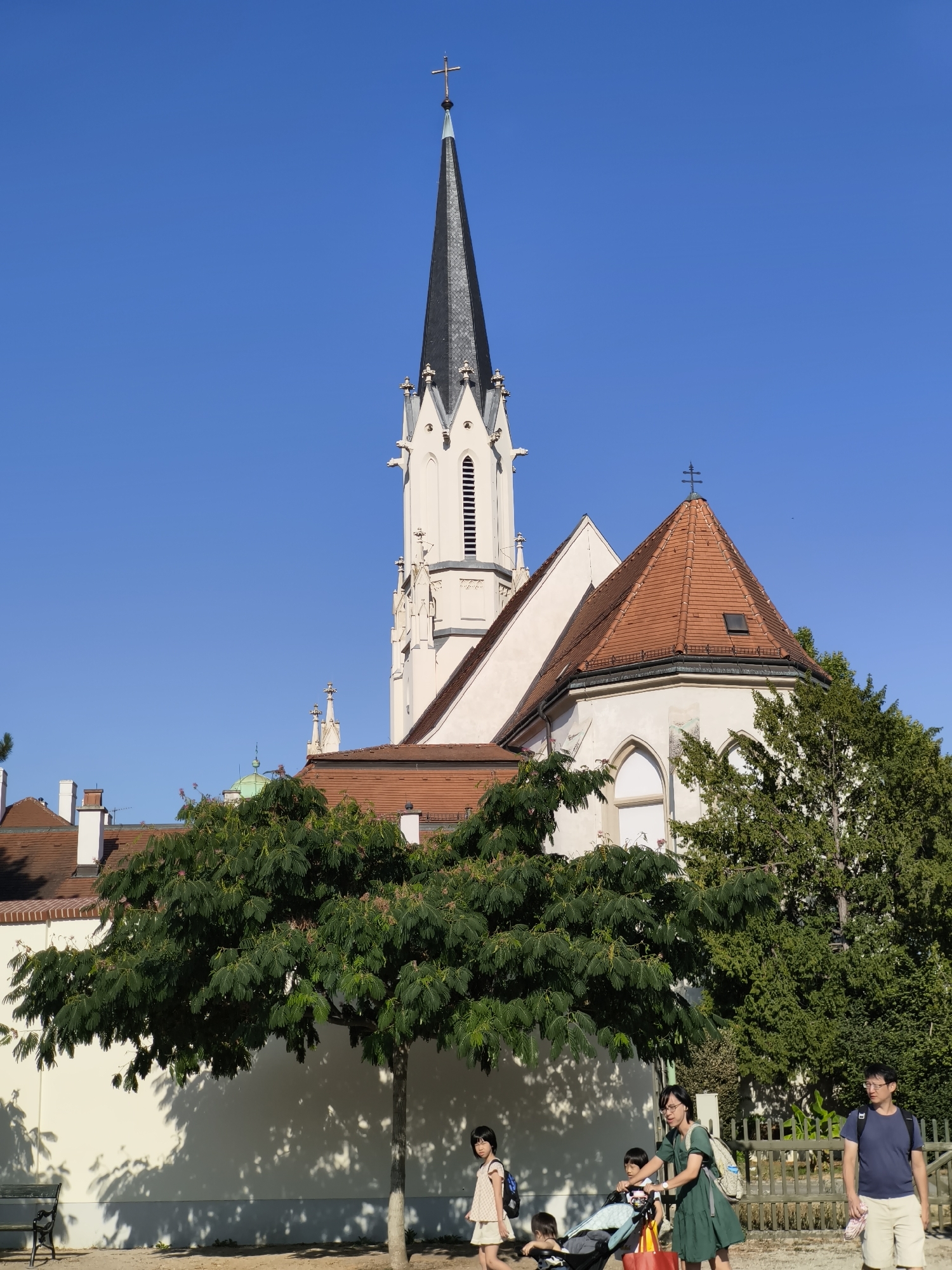 In Vienna, Austria, a tall, pale church with a pointed black spire dominates the background under a clear blue sky. A portion of the church's red-tiled roof is visible, adorned with a cross. In the foreground, a family is seen with a stroller and children. A woman in a green dress pushes the stroller, accompanied by two children. Another child stands to her left, and a man in a blue shirt and shorts walks to the right. A lush green tree with delicate foliage stands prominently in the middle ground, casting dappled shadows on a white wall behind it. A park bench is visible to the left. The overall scene depicts a bright, sunny day with people enjoying an outdoor space near a historic church.