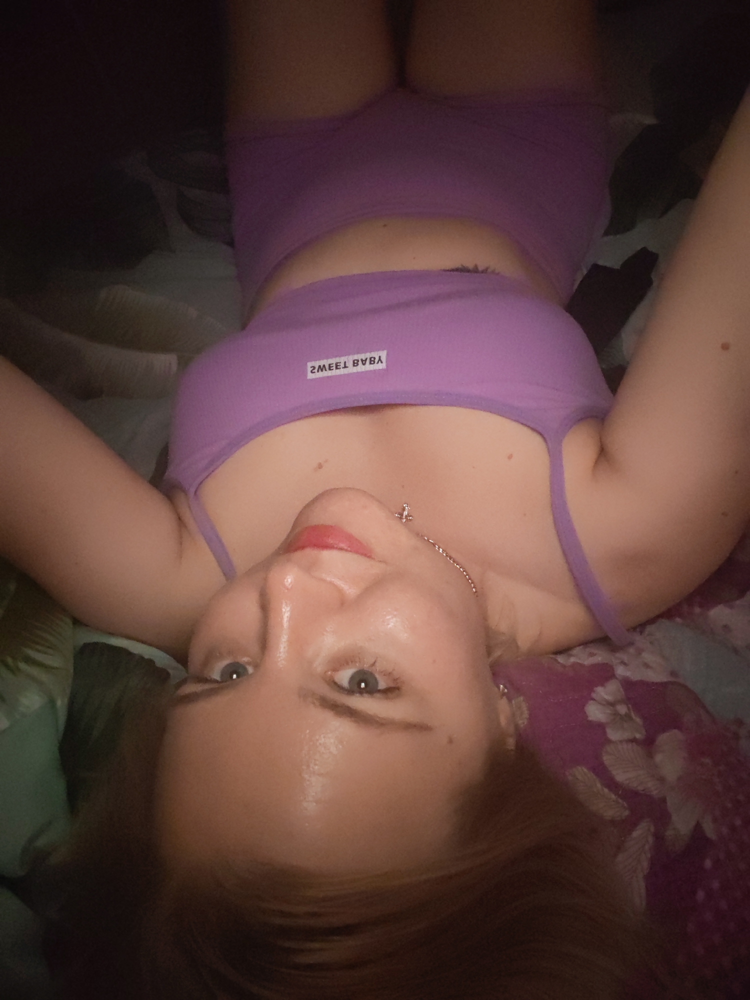 A woman with fair skin and blue eyes is lying on a bed, wearing a lavender-colored crop top and shorts. She is positioned with her head at the bottom of the frame and her torso towards the top. The bed has a floral patterned bedspread. The lighting is subdued. There is no information in the image to indicate a location in the city of Izhevsk, Russia.