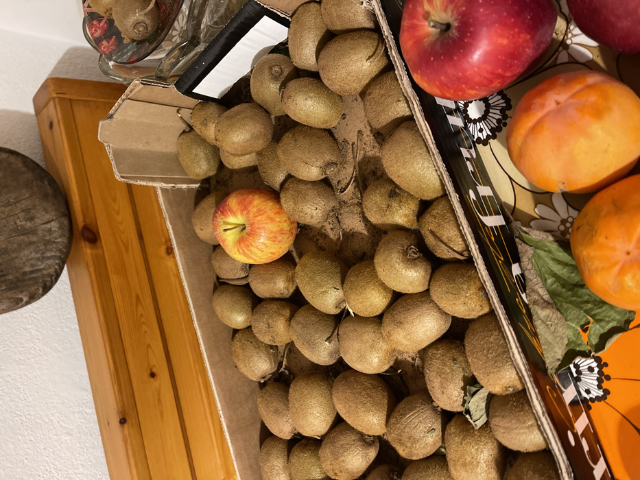 This media file captures an indoor setting in Torre di Santa Maria, Italy, focusing on a collection of fruits. The primary subjects are a large quantity of kiwis, two apples, and two persimmons.

A substantial cardboard box, partially open and with some visible dirt/fibers inside, holds numerous kiwis, appearing to be freshly harvested or recently acquired. Nestled among these kiwis is one small, round apple with red and yellow stripes. To the right of the box, another larger, deep red apple is visible. Alongside this apple are two ripe, orange persimmons, resting on a patterned surface that features floral or geometric designs in shades of cream and brown. A dark green leaf is also present near one of the persimmons.

The box and surrounding fruits are placed on a warm-toned wooden surface, likely a table or countertop. To the left, a portion of a round, rustic wooden object, possibly a cutting board, is visible, alongside a white wall in the background. The lighting suggests an ambient indoor environment.

The side of the cardboard box displays an unreadable black and white logo or text. No people are visible, and no specific activity is taking place beyond the arrangement or storage of the fruits. The scene evokes a sense of home or a local market display.