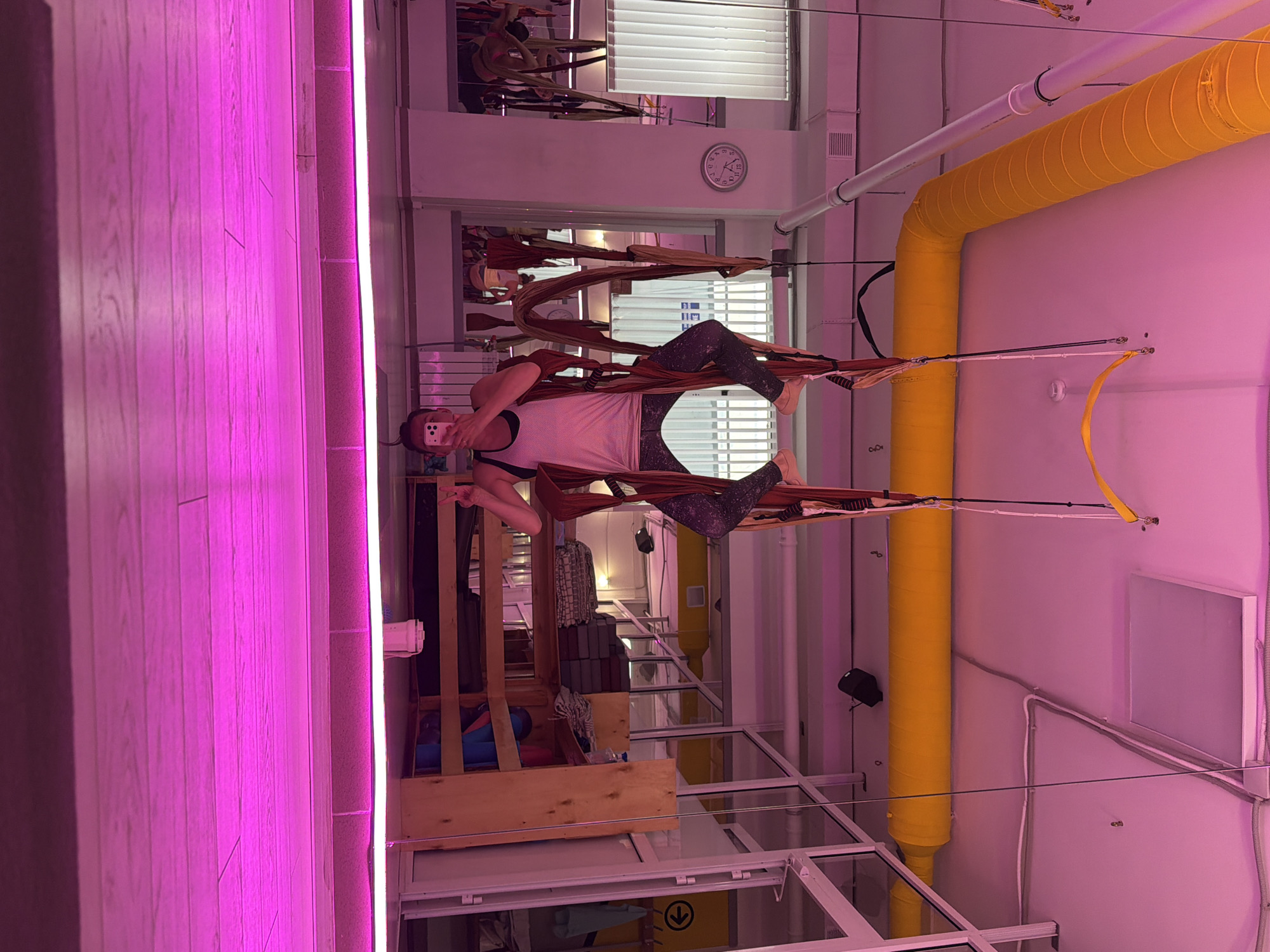 An inverted person is engaged in aerial yoga within a studio. The individual is suspended upside down, supported by a hammock. The environment is characterized by a pinkish-purple hue, likely from artificial lighting, illuminating the wooden floor and a raised platform. Yellow pipes are visible along the ceiling, and other yoga equipment, such as mats and bolsters, are arranged on shelves in the background. Mirrors line one wall, reflecting the studio space. The location is identified as Chaikovsky, Russia.
