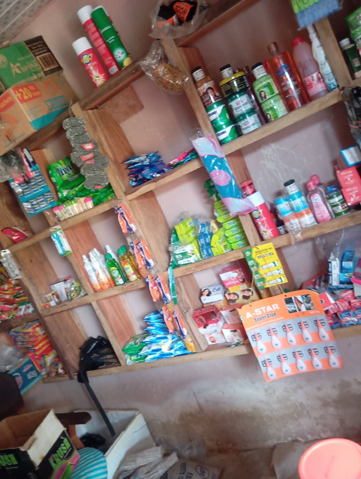 The image depicts the interior of a small shop or stall, likely a convenience store or a small retail establishment, located in Jalingo, Nigeria. The shop is filled with various consumer goods stacked on wooden shelves that are informally constructed.
The shelves are laden with a variety of products, including cleaning supplies, toiletries, food items, and possibly some stationery. Visible items include:
* **Top Shelf:** Aerosol cans (possibly insect repellent or air fresheners) in red and green packaging, a bag of what appears to be rubber bands or similar small elastic items, and other miscellaneous packaged goods.
* **Middle Shelves:** Rows of small packaged items, possibly soaps or disinfectants, in green and white packaging. There are also bottles of liquids, some appearing to be oils or medicinal concoctions, in various colors. Packets of what look like tissues or wet wipes are also present. A display of toothpaste or similar personal care items is visible. A prominent display card for "A-STAR Super Glue" with multiple tubes of glue is attached to one of the shelves.
* **Lower Shelves and Floor:** More packaged goods, including what appear to be snacks or biscuits in brightly colored wrappers. Cardboard boxes are stacked, one labeled with "40g". A black plastic bag is positioned on a shelf. Towards the bottom of the image, there are cardboard boxes on the floor, one displaying the word "KRUSH". A light blue and white striped item is also on the floor.
The lighting is somewhat dim, and the image quality is moderate, suggesting it was taken in low light conditions or with a basic camera. The overall impression is of a cluttered but functional retail space catering to everyday needs. The environment appears to be an indoor setting, likely a small shop in a commercial or residential area of Jalingo. There are no people clearly visible in the image.