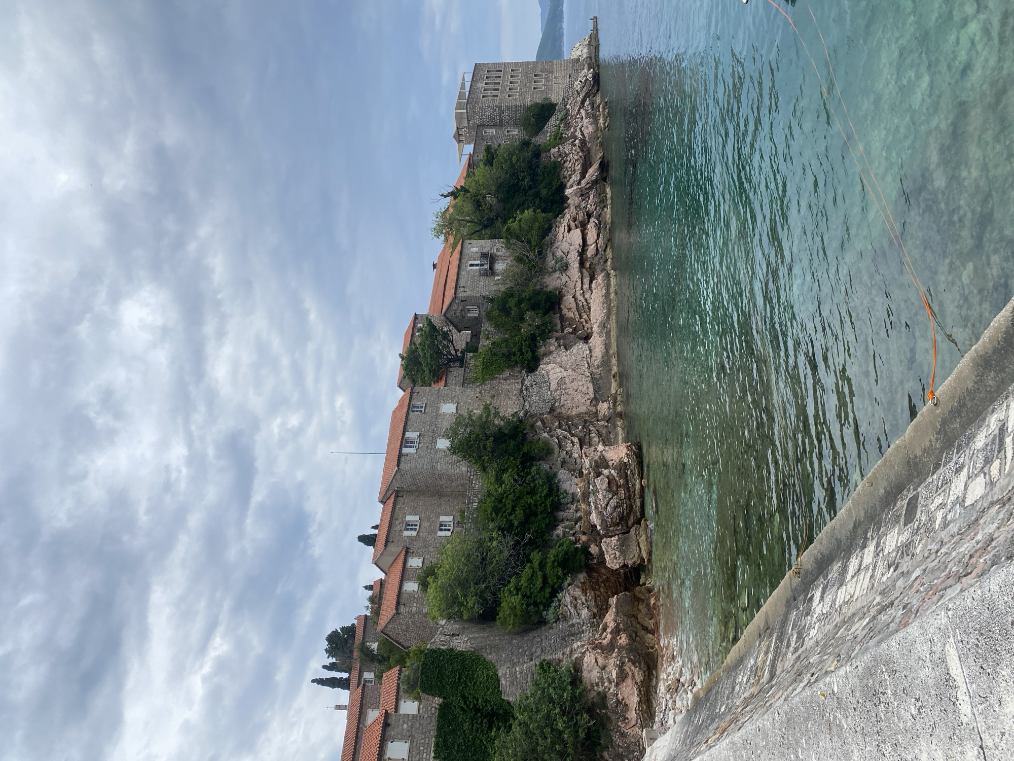 The image captures a view of Sveti Stefan, Montenegro. Buildings with red-tiled roofs are visible atop a rocky coastline that descends into clear, turquoise waters. The structures appear to be historical and are surrounded by green vegetation. The sky is overcast, with clouds filling the upper portion of the image. A stone wall runs along the bottom of the image, suggesting a pathway or edge of a structure. An orange rope is visible in the water.