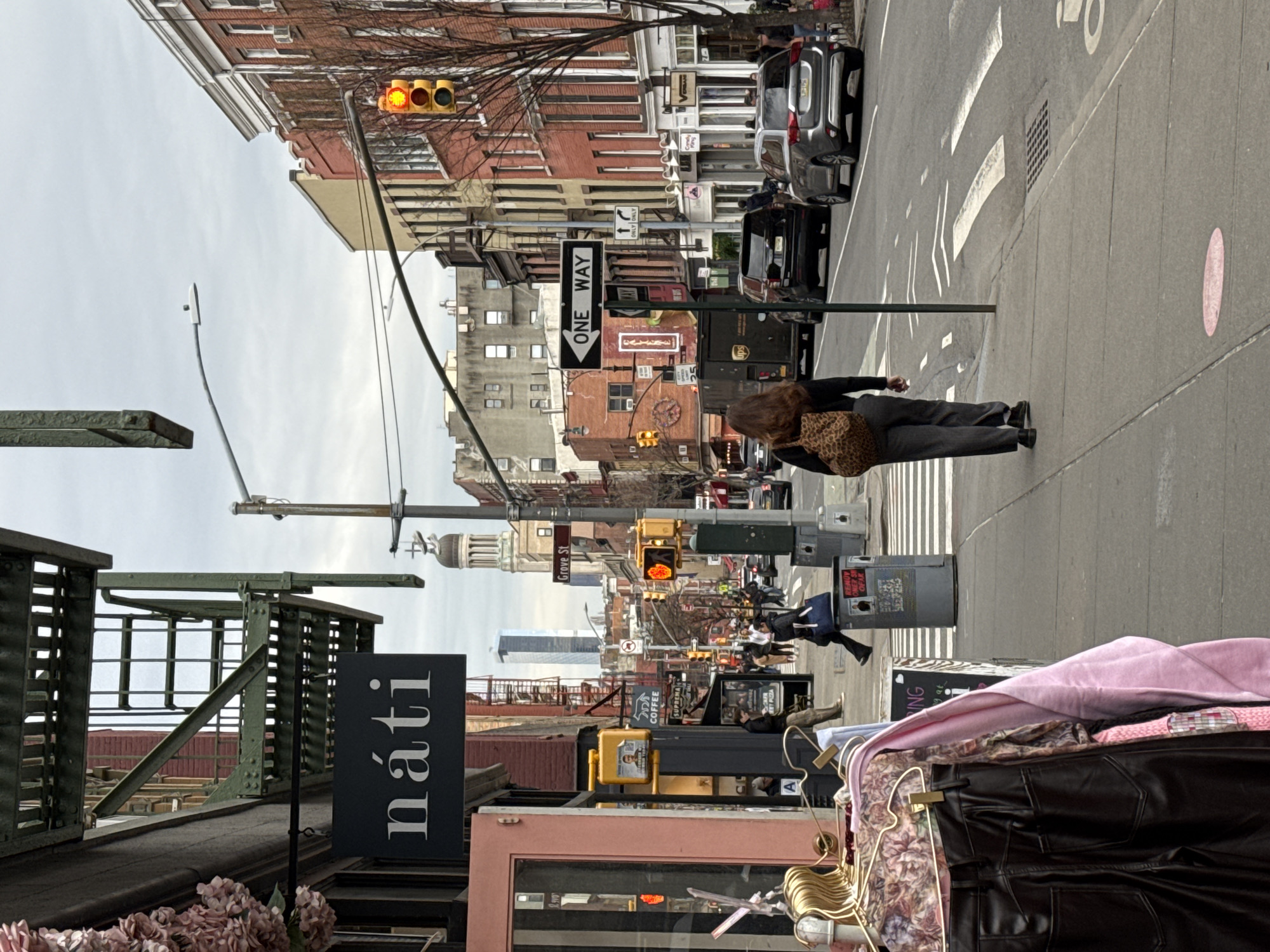 This image depicts a street scene in Greenwich Village, Manhattan, New York City, United States of America, looking down a street with the Jefferson Market Library visible in the distance. The sky is overcast.
In the immediate foreground on the left, a display of clothing on gold hangers includes black faux leather pants, a patterned blouse, and a pink long-sleeved garment. Above this, a black rectangular sign with "náti" in white serif lettering is suspended. Artificial pink floral arrangements are partially visible in the upper left. A dark green metal fire escape is attached to the brick building.
On the sidewalk, a woman with long dark brown hair, wearing a black top and grey trousers, walks away from the camera, carrying a leopard-print shoulder bag on her right shoulder. A grey cylindrical trash receptacle is located near a pedestrian crossing. Further along the sidewalk, other pedestrians are visible.
The street features a pedestrian crosswalk and designated bike lanes. A utility pole holds multiple street signs, including a "ONE WAY" sign with a right-pointing arrow and a "Grove St" sign. Traffic lights are visible, with red signals facing the viewer's direction of travel. Vehicles on the street include a dark brown UPS delivery truck, a black SUV, and a grey SUV.
The street is lined with multi-story buildings, primarily constructed of brick in various hues of red, orange, and light yellow. Many buildings exhibit classic New York fire escapes and visible air conditioning units. In the mid-background, the domed tower of the Jefferson Market Library is a prominent architectural feature. Taller, modern skyscrapers are discernible in the far background, indicating the broader urban skyline.