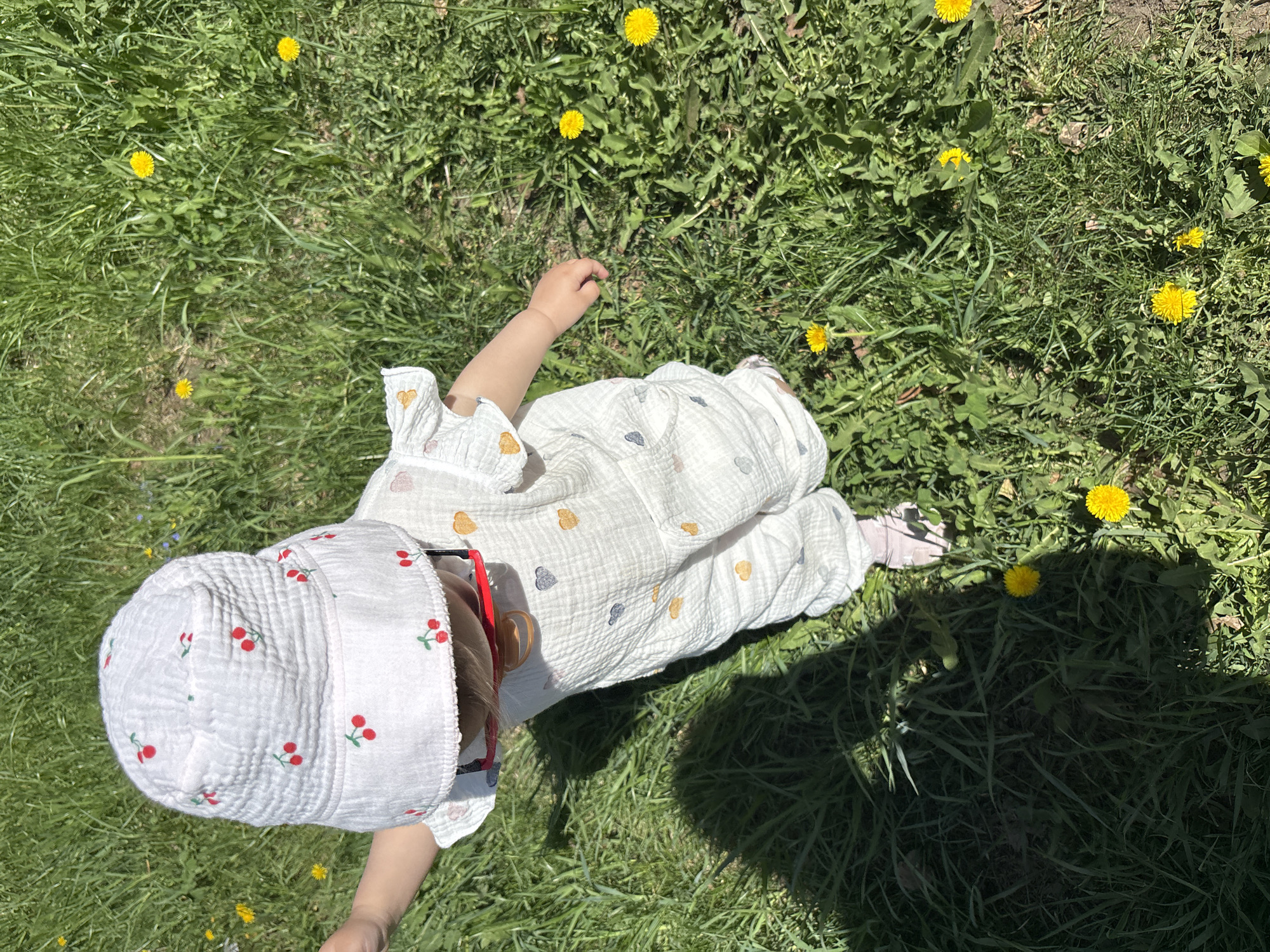 A baby wearing a white cherry-patterned hat and a white onesie with heart patterns lies on a patch of green grass dotted with dandelions. The location appears to be outdoors, likely a park or lawn. The child's face is partially obscured by the hat. The photo is taken from a high angle.