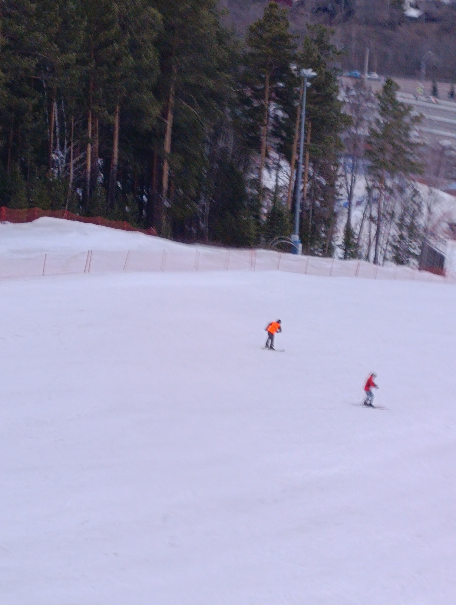 The image depicts a winter scene on a snow-covered slope, likely a ski hill, with two individuals skiing downhill. The person closer to the top of the image is wearing an orange jacket. The person closer to the bottom of the image is wearing a red jacket. The slope is surrounded by a sparse barrier fence, with a backdrop of evergreen trees and what appears to be a city or highway visible in the distance. Based on the question context, the location is likely a ski area in or near the city of Krasnoyarsk, Russia.