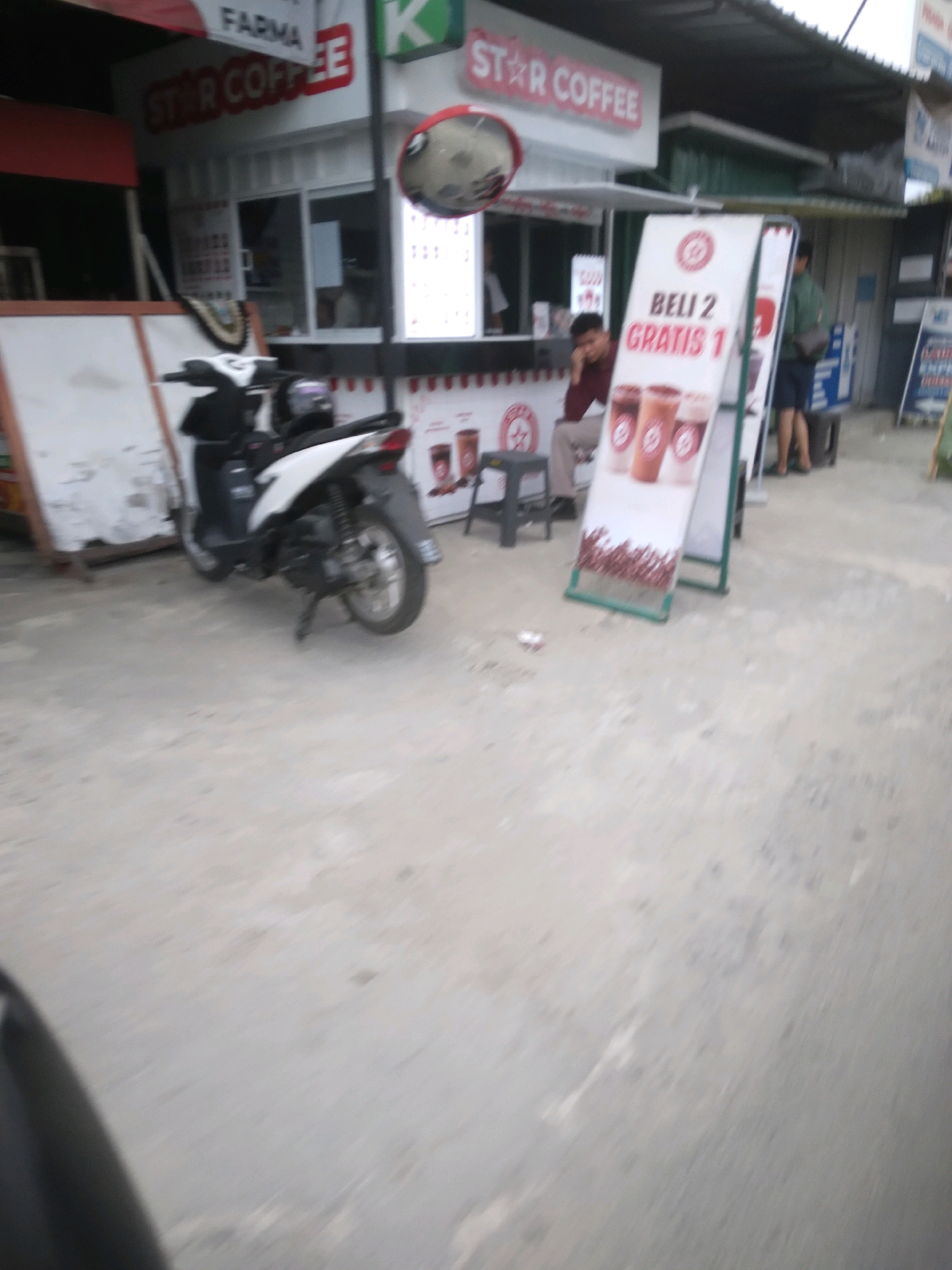 The image depicts a street scene in Desa Pamijen, Kecamatan Sokaraja, Indonesia, outside a "STAR COFFEE" establishment. A white scooter is parked on the left side of the frame. In front of the coffee shop, a man is seated on a dark stool, leaning forward. A prominent A-frame sign is positioned in front of the coffee shop, displaying text in Indonesian, "BELI 2 GRATIS 1," along with images of three coffee drinks. Another person, possibly an employee, is standing behind the sign, and another individual is seen in the background near a blue stand. The overall environment appears to be an outdoor commercial area with a concrete ground surface.