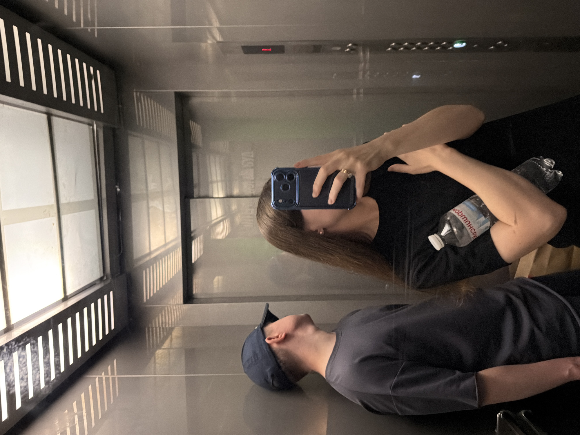 In an elevator in Kyiv, Ukraine, a person wearing a dark blue baseball cap and a dark grey t-shirt leans against the wall, looking upwards. Beside them, another person, with long brown hair, holds a clear plastic bottle of water in their left hand and a smartphone in their right, using it to take a mirror selfie. The elevator has metallic walls and a ceiling with linear lighting fixtures that cast striped shadows. A red light is illuminated on the elevator control panel.