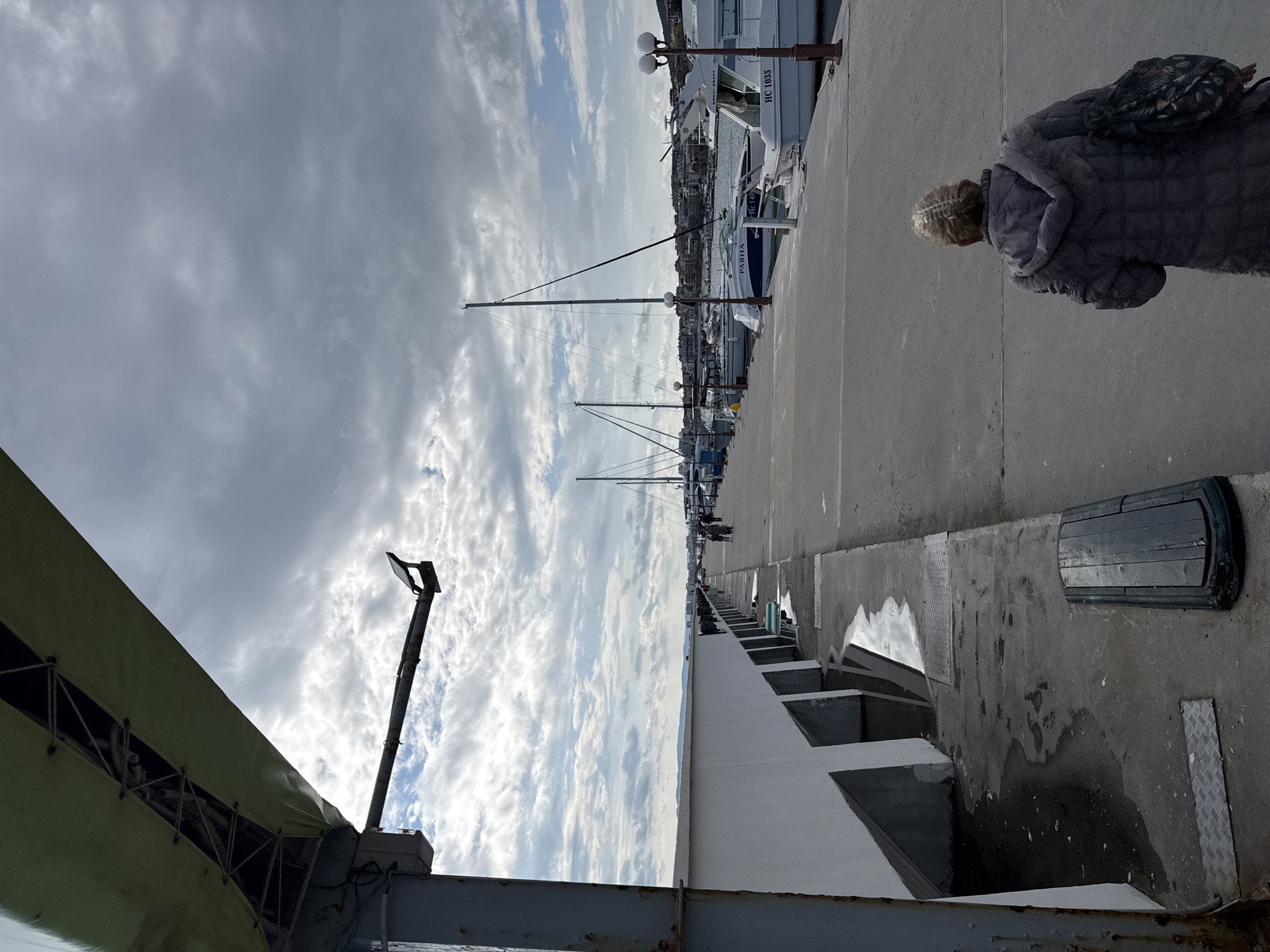The image depicts a pier or harbor setting in Sveti Vlas, Bulgaria, under a sky filled with thick clouds. In the foreground, part of a metallic structure with a green awning is visible on the left. The pier extends into the distance, lined with boats and sailboats docked on one side. Several masts are visible against the cloudy sky. A woman wearing a grey coat and carrying a patterned bag walks on the pier. The pier's edge is marked by a white structure with geometrical, step-like divisions. Puddles and damp patches indicate a recent rain or wet conditions. Other people can be seen farther down the pier.