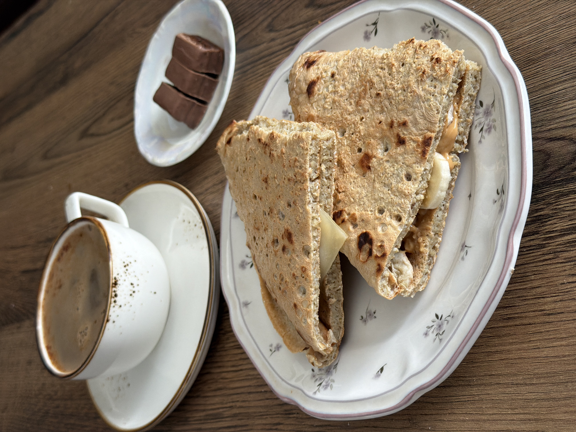 A still life on a wooden surface features a cup of coffee on a saucer, a small dish of chocolate pieces, and a plate containing two halves of a triangular sandwich. The sandwich appears to have a filling of peanut butter, banana slices, and cheese. The scene's location is undetermined as the image lacks context and discernible landmarks that would indicate a location in Perm, Russia, or any other city.