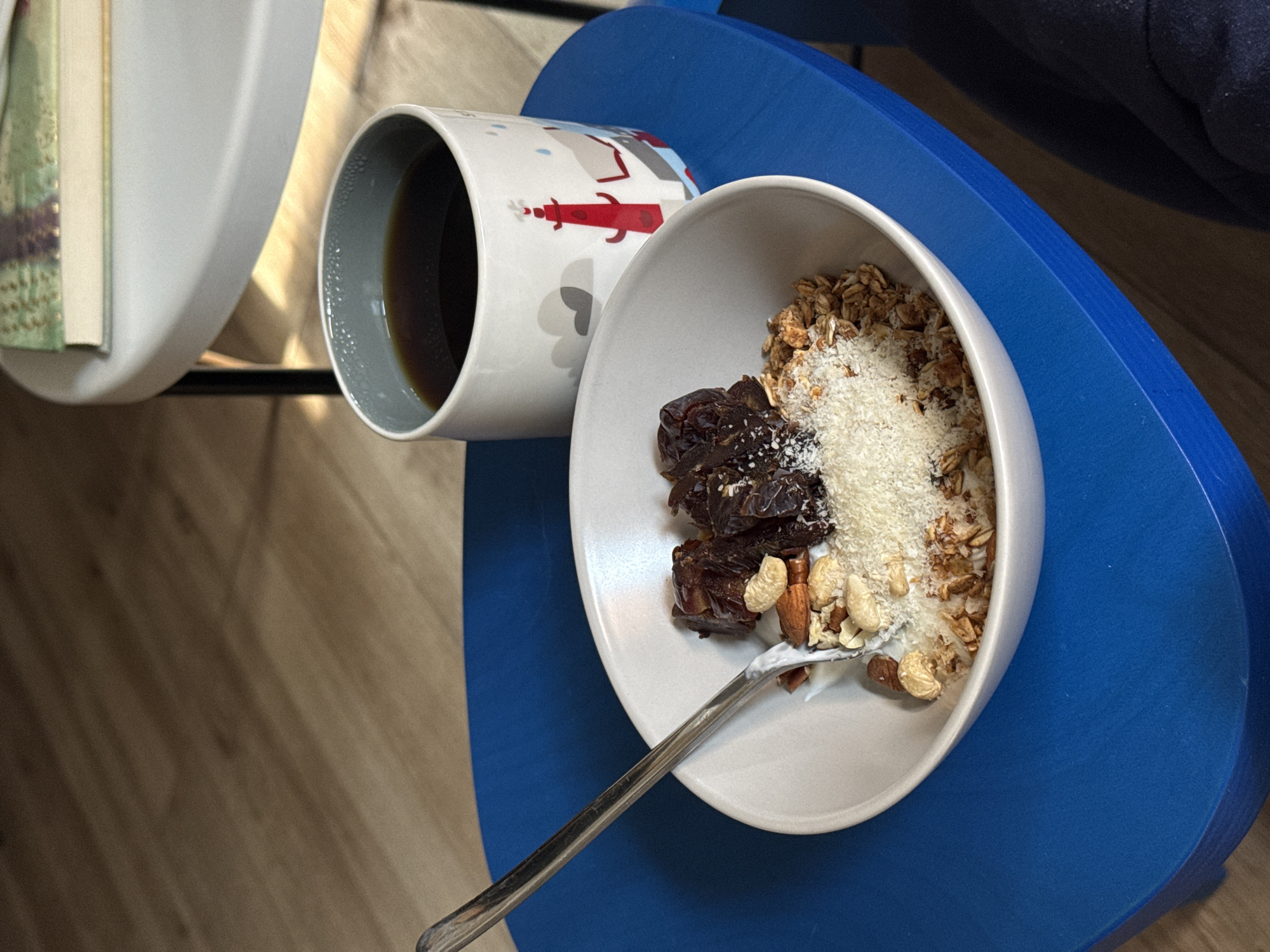 A close-up shot captures a bowl of granola topped with dates, almonds, and shredded coconut, alongside a mug of black coffee. A silver spoon rests in the bowl, indicating someone is about to eat. The scene is set on a bright blue table, with a glimpse of a book on a white side table in the background. The overall impression is one of a cozy breakfast setup, possibly in a cafe or home in Tel-Aviv, Israel. The lighting suggests a natural light source, possibly from a window, creating a warm and inviting atmosphere.