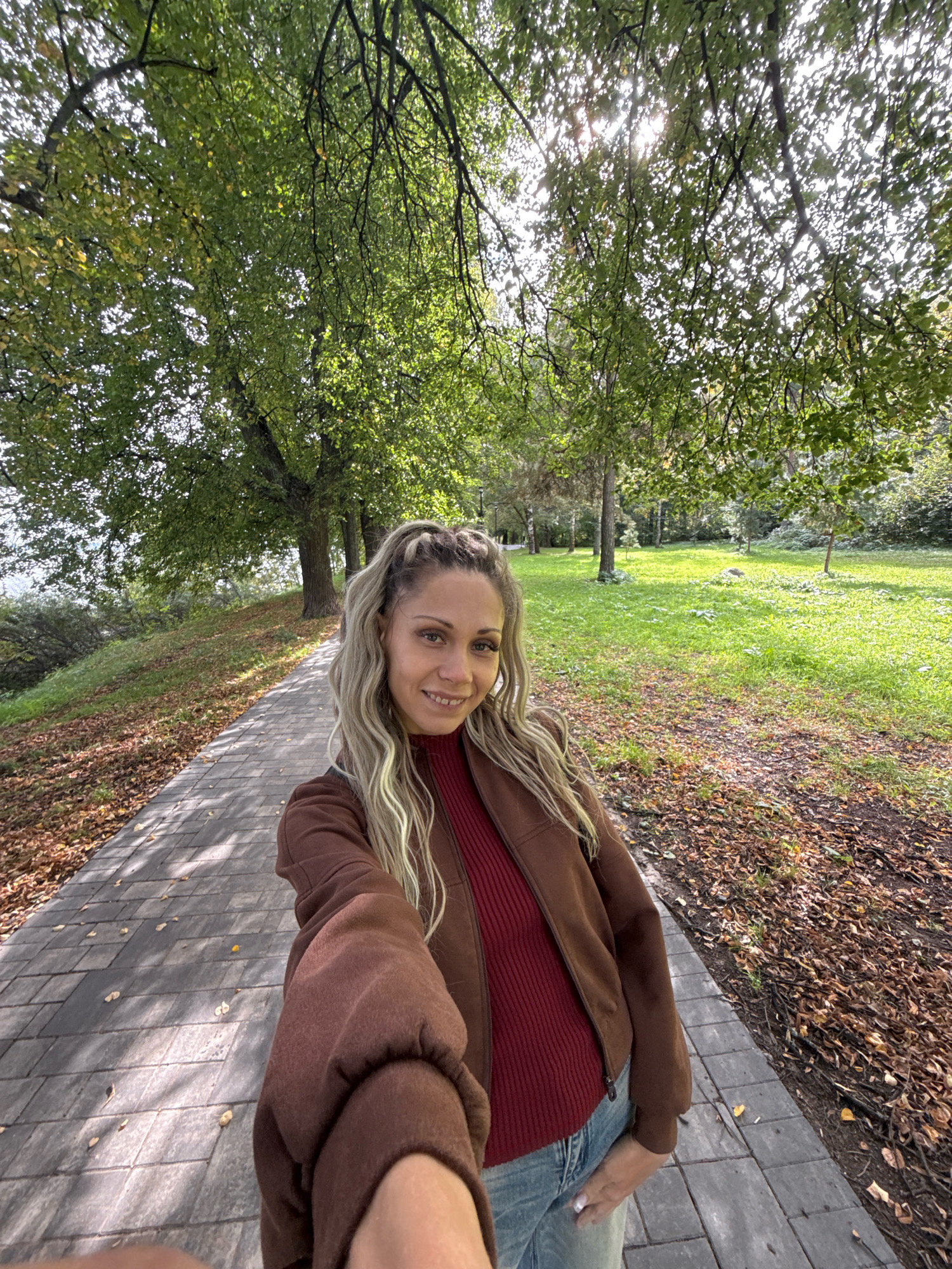 A woman with blonde hair braided and pulled to one side, is taking a selfie in a park in Zhukovo, Russia. She is wearing a maroon ribbed sweater and a brown zip-up jacket, and blue jeans. She is smiling at the camera. The park has a paved walkway with fallen leaves scattered on it. Tall trees with green leaves line the walkway, and a grassy area is visible to the right. The sun is shining through the trees, creating shadows on the ground.