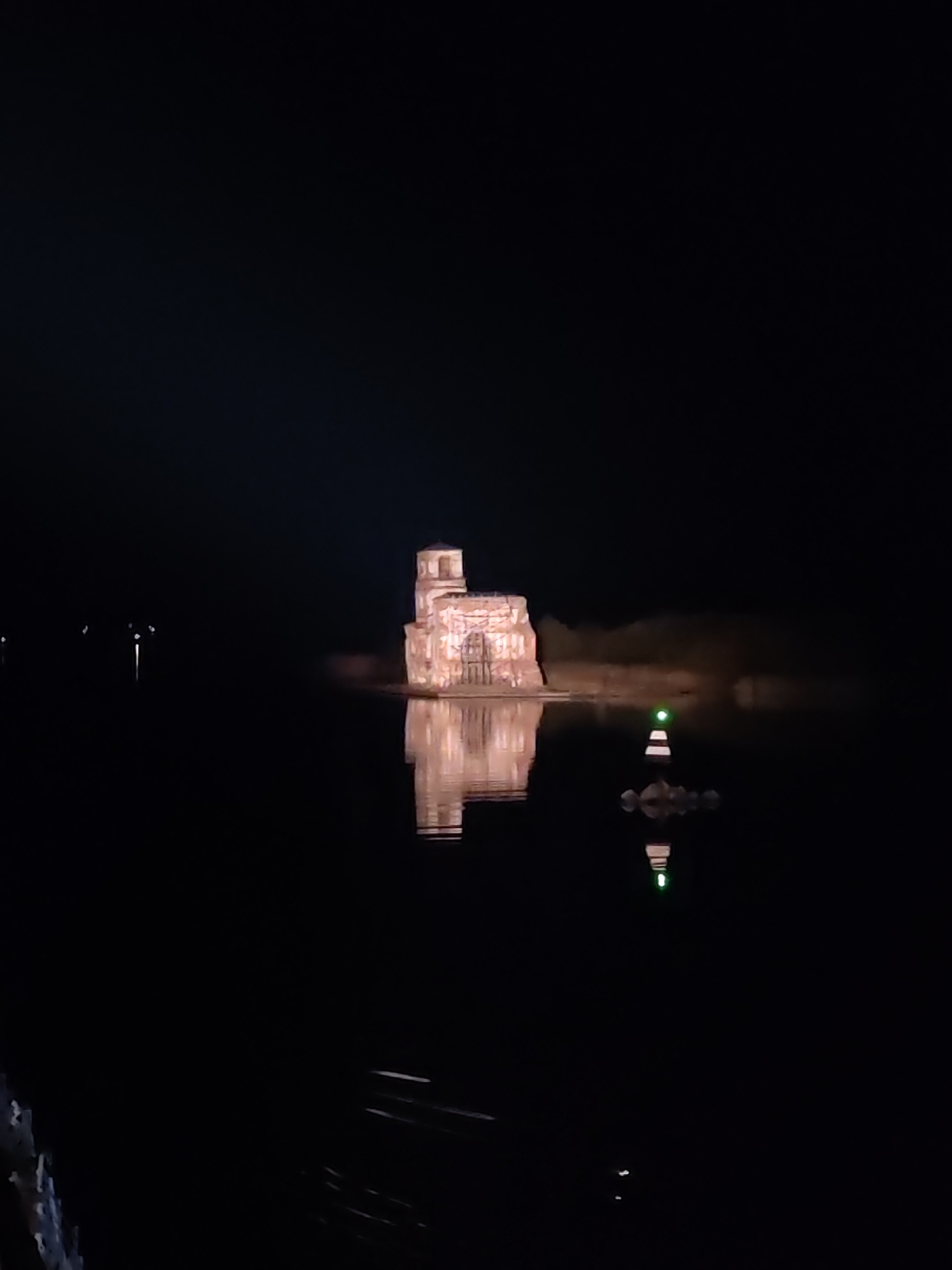 An illuminated architectural structure, likely a historical building, stands in water under a dark sky. The structure appears to have multiple levels and is lit with a warm glow, with its reflection visible in the water. To the right of the structure is a small, striped lighthouse or marker with a green light on top, also reflected in the water. The scene appears to be at night, and the surrounding environment is dark with very little ambient light. The location is Krokhino, Russia.