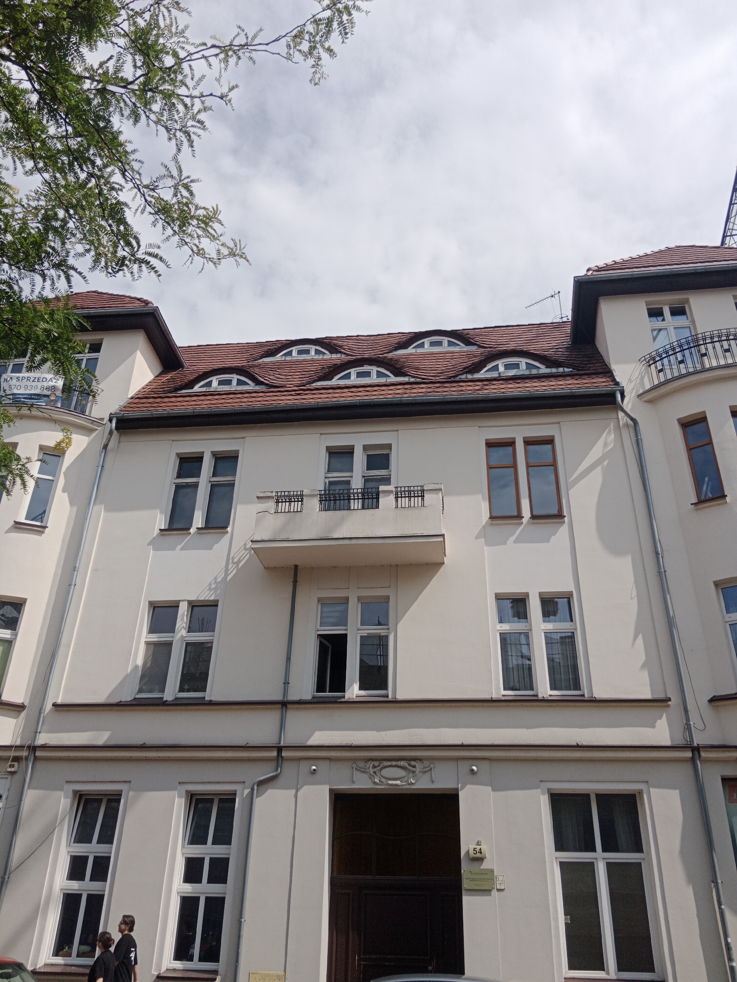 This image depicts the facade of a cream-colored, multi-story building with a red tile roof. The building features several windows of various sizes and styles, including some with arched tops integrated into the roofline. A balcony with a black metal railing is visible on one of the upper floors. In front of the building, there are two individuals, one appearing to be a younger person, both wearing dark clothing. The lower part of the image shows a dark wooden entrance door with a plaque labeled "54". A portion of a green tree with dense foliage is visible in the upper left corner, partially obscuring the sky and part of the building. The scene appears to be on a street in Bydgoszcz, Poland.