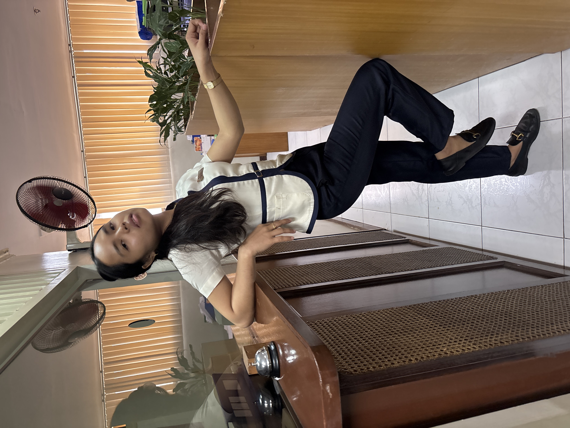 A young woman with dark hair pulled back stands next to a counter. She is wearing a white collared shirt with navy blue trim and navy blue pants. She has a watch on her left wrist and a ring on her right ring finger. Her left arm is resting on the counter, and her right leg is bent with her foot resting on the counter. The counter is made of dark wood and has a bell on it. Behind the counter are some plants and blinds. There are two fans on the ceiling. The floor is tiled. The scene appears to be an indoor reception area, possibly in a hotel or office building. The specific location in Davao City, Philippines, is not provided in the image.