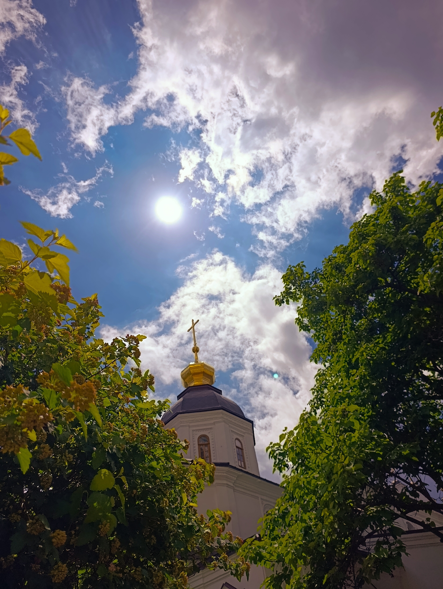 This is a low-angle, close-up shot of a golden-domed church tower topped with a cross, partially obscured by lush green trees and foliage. The sky above is a bright blue with scattered white clouds, and the sun is a bright, glowing orb. The architectural style of the church, with its white facade and arched windows, suggests it is likely an Orthodox church, common in Kyiv, Ukraine. The vibrant greenery indicates a warm season, likely spring or summer. The overall impression is one of peace and natural beauty set against an architectural backdrop.