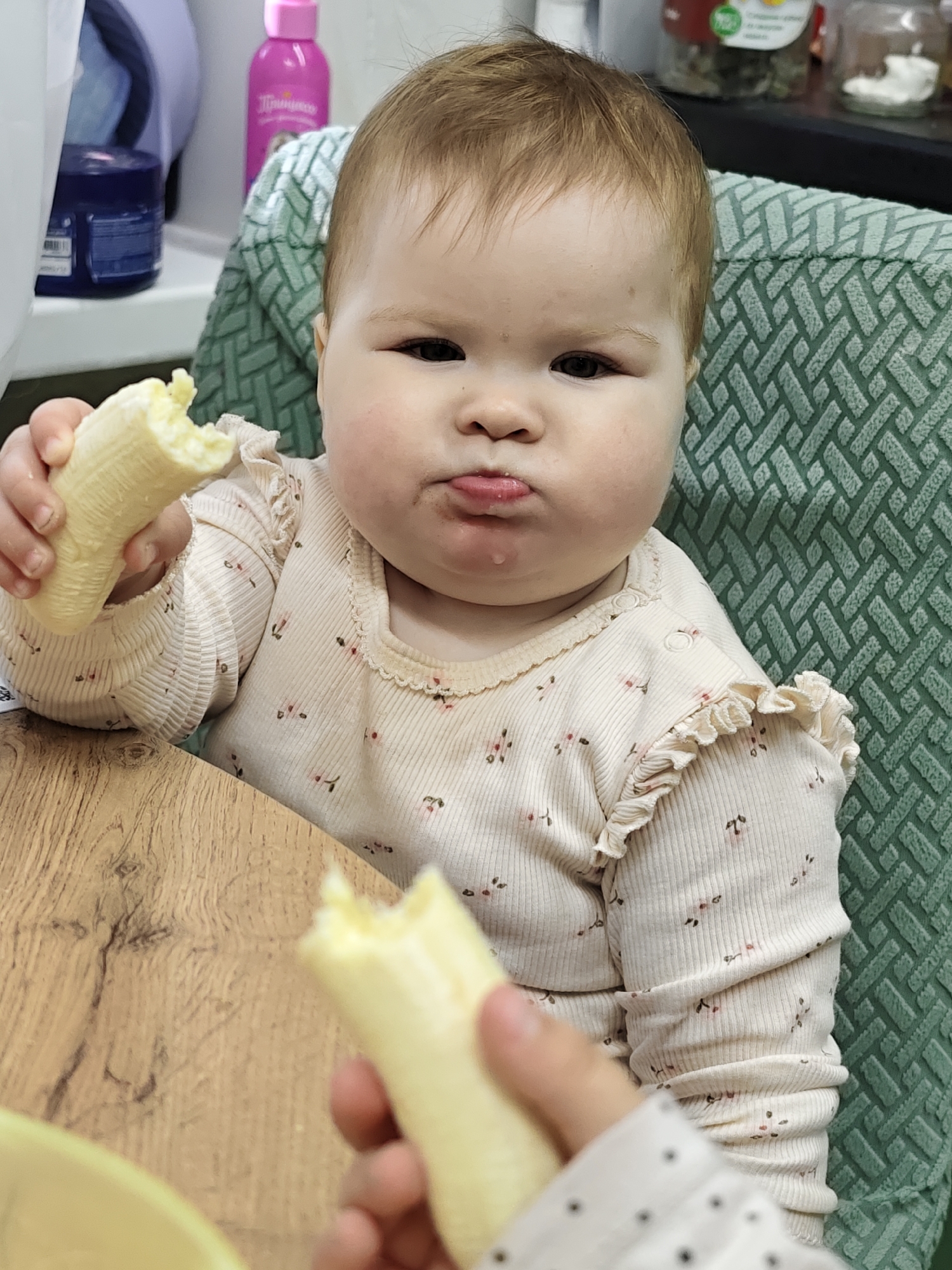 A baby with light brown hair and chubby cheeks sits in a high chair with a green patterned fabric. The baby is wearing a light-colored, long-sleeved shirt with small floral designs and ruffled edges. They are holding a partially eaten banana in each hand, and a drop of liquid hangs from their pursed lips. The baby appears to be indoors. In the background, there are several containers on a shelf. A wooden-looking table is in the foreground. The location is Chaikovsky, Russia.
