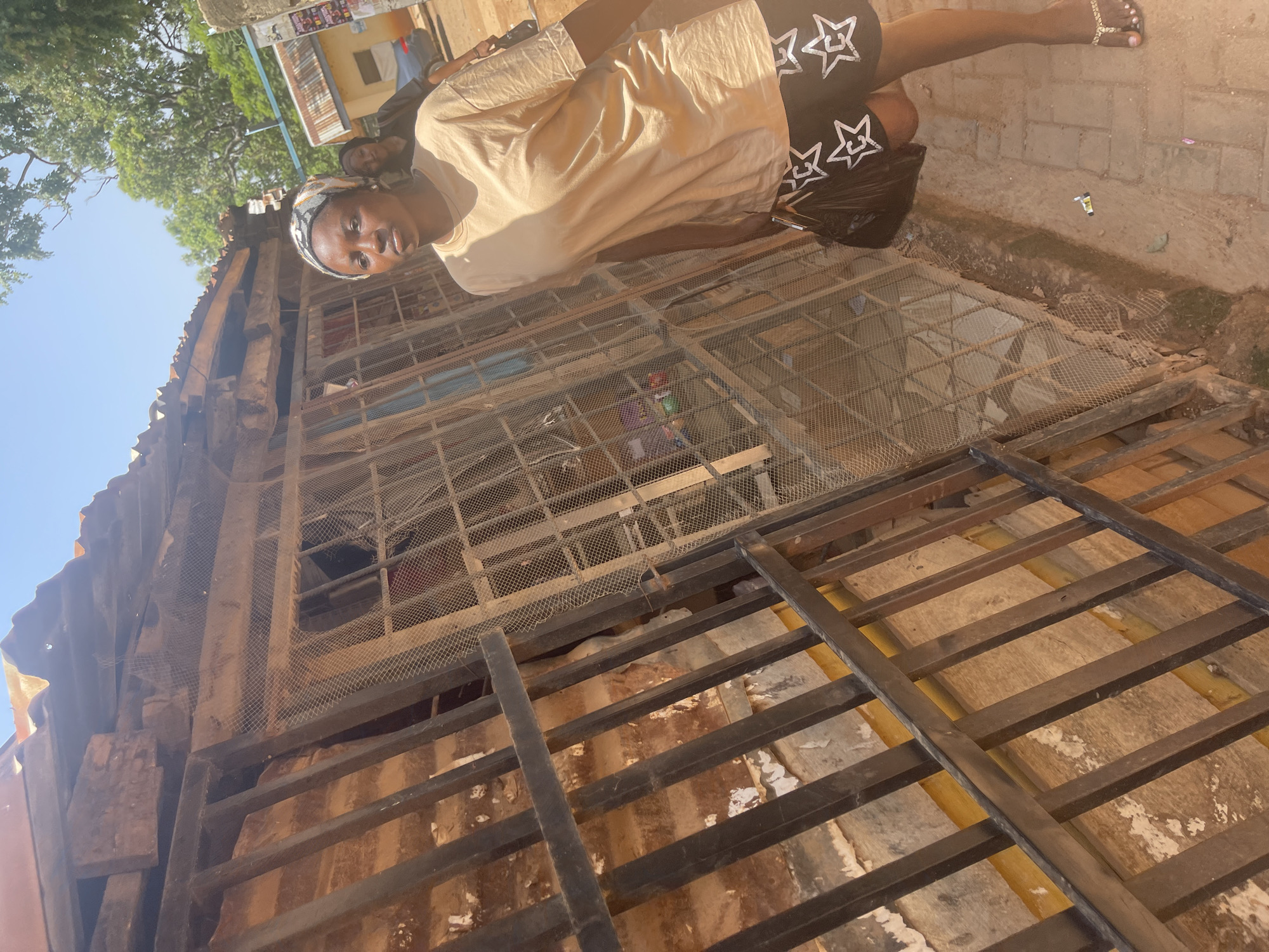 A young Black woman is positioned in the foreground of the image, facing sideways and looking towards the camera. She wears a light-colored, loose-fitting t-shirt and black shorts adorned with white star patterns. A black plastic bag is held in her left hand, and she is wearing sandals. Her expression appears neutral. She stands near a rustic, weathered structure with an open, caged-like facade, possibly a market stall or an unfinished building, made of metal bars and wire mesh. The background shows more of this structure and a hint of trees and a bright blue sky.
The scene appears to be outdoors, likely in a somewhat informal or developing area, given the construction materials and the natural elements visible. The lighting suggests it is daytime, with bright sunlight casting shadows. The overall atmosphere is casual and everyday. There is no discernible text visible in the image. The image is oriented vertically, and the scene is captured from a slightly elevated perspective looking down.