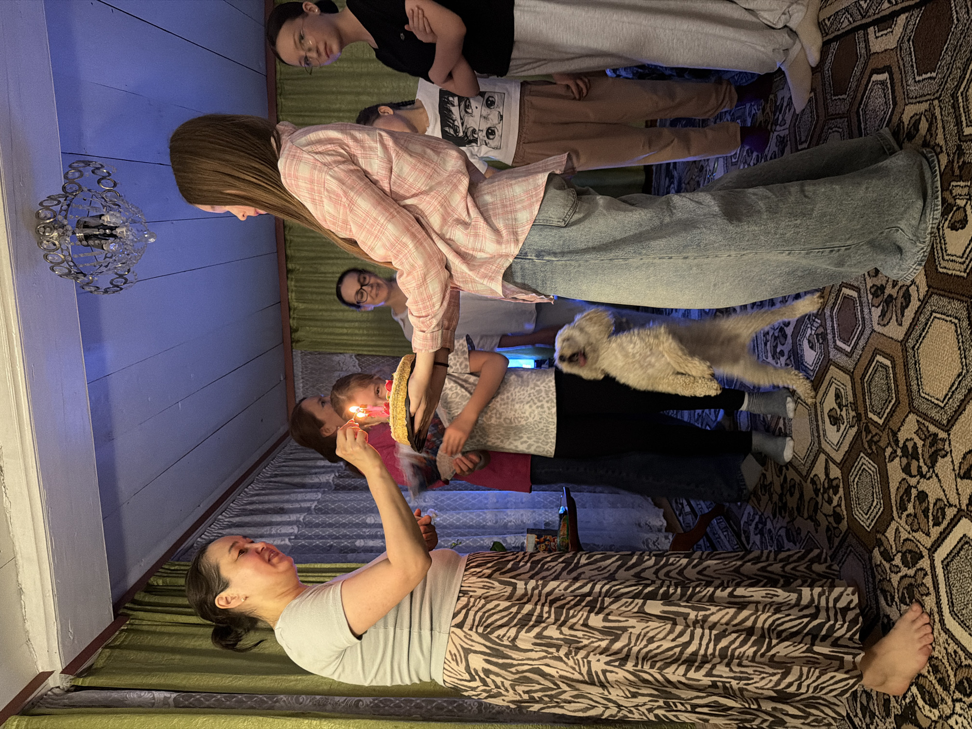 The image depicts a group of people gathered indoors, likely celebrating a birthday. A woman holds a cake with lit candles, and a child is close to her. Several other individuals are observing the scene. A dog is in mid-air, possibly jumping. The room has a patterned rug and a light fixture hanging from the ceiling. Based on the prompt, the location is Кадряково, Russia.