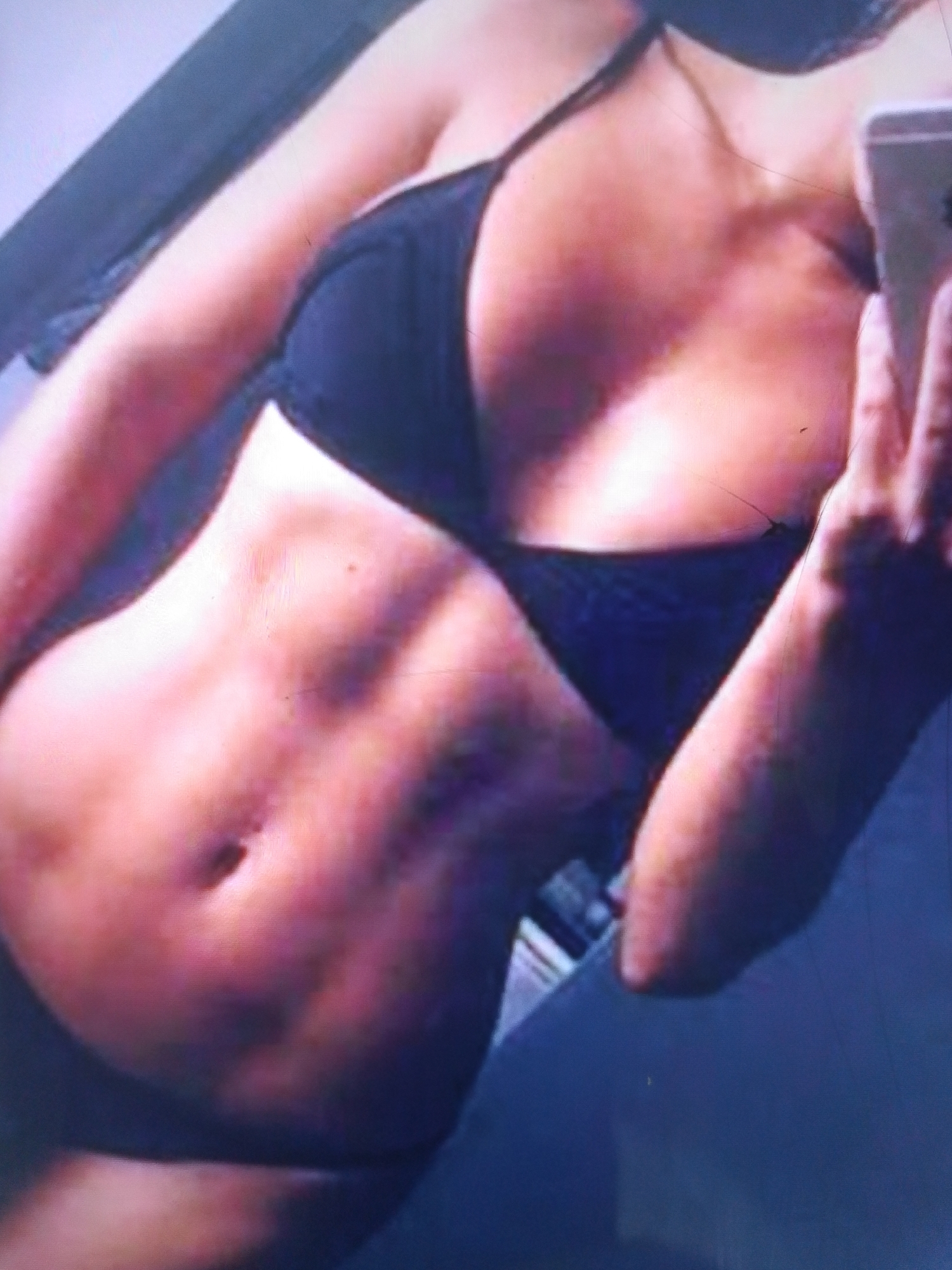 The image shows a woman posing in swimwear, holding a smartphone. She has visible abdominal muscles and is wearing a black bikini top and bottom. The lighting suggests an indoor setting, potentially a bedroom or changing room. The background is out of focus, but appears to be a wall or furniture. There is no discernible information in the image that would place it in Dobra Krynytsia, Ukraine, or any other specific urban location.