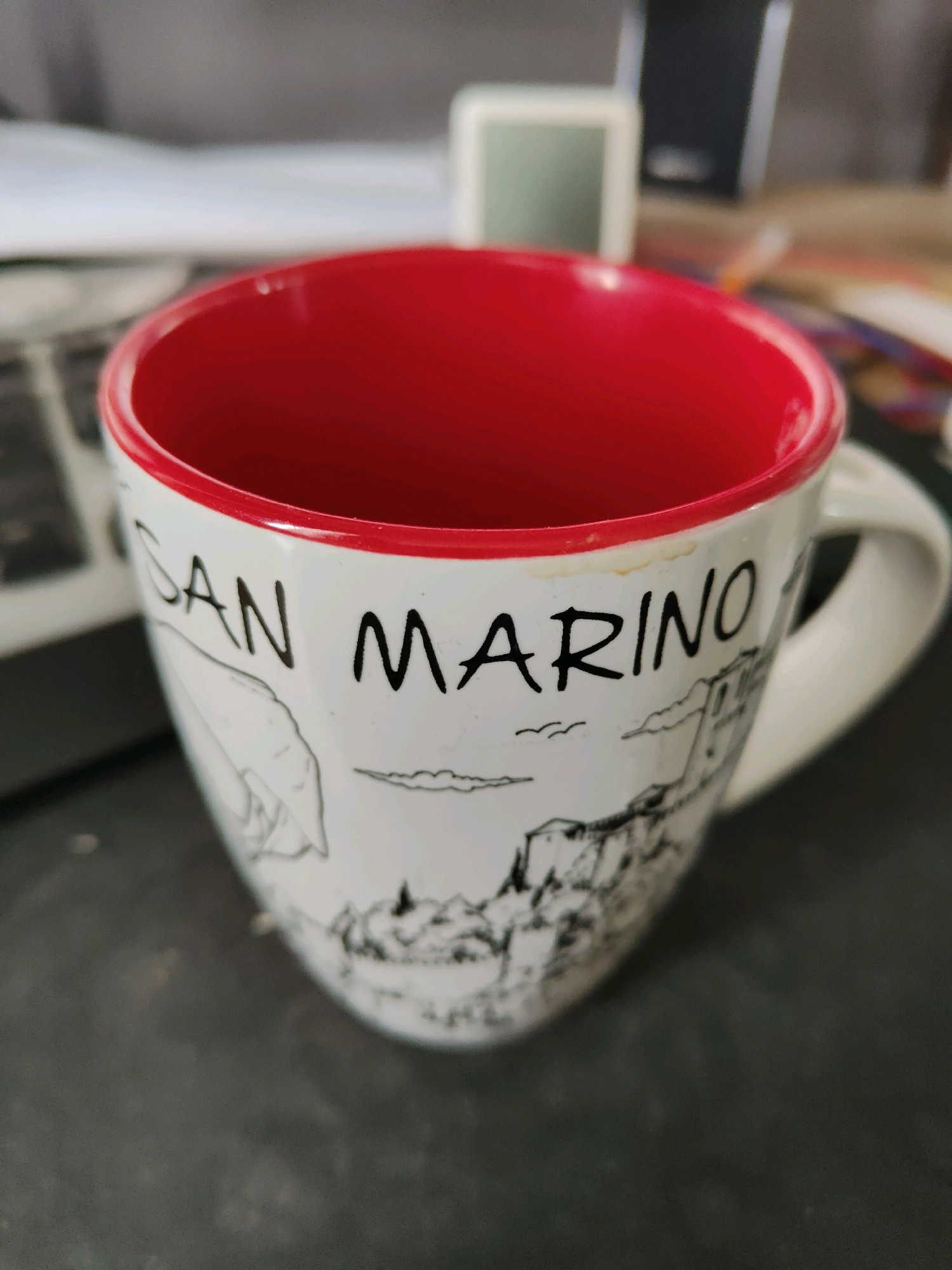 A close-up, eye-level shot shows a white ceramic mug with a red interior. The exterior of the mug is decorated with a black line drawing of buildings and landscape, and the words "SAN MARINO" are printed in black lettering. A handle is visible on the right side of the mug. The mug is positioned on a dark gray, textured surface, possibly a desk. The background is blurred, showing a hint of electronic devices and a light-colored object. The image appears to be taken indoors. There is no identifiable information relating to the city of Homyel, Belarus, or any specific location.