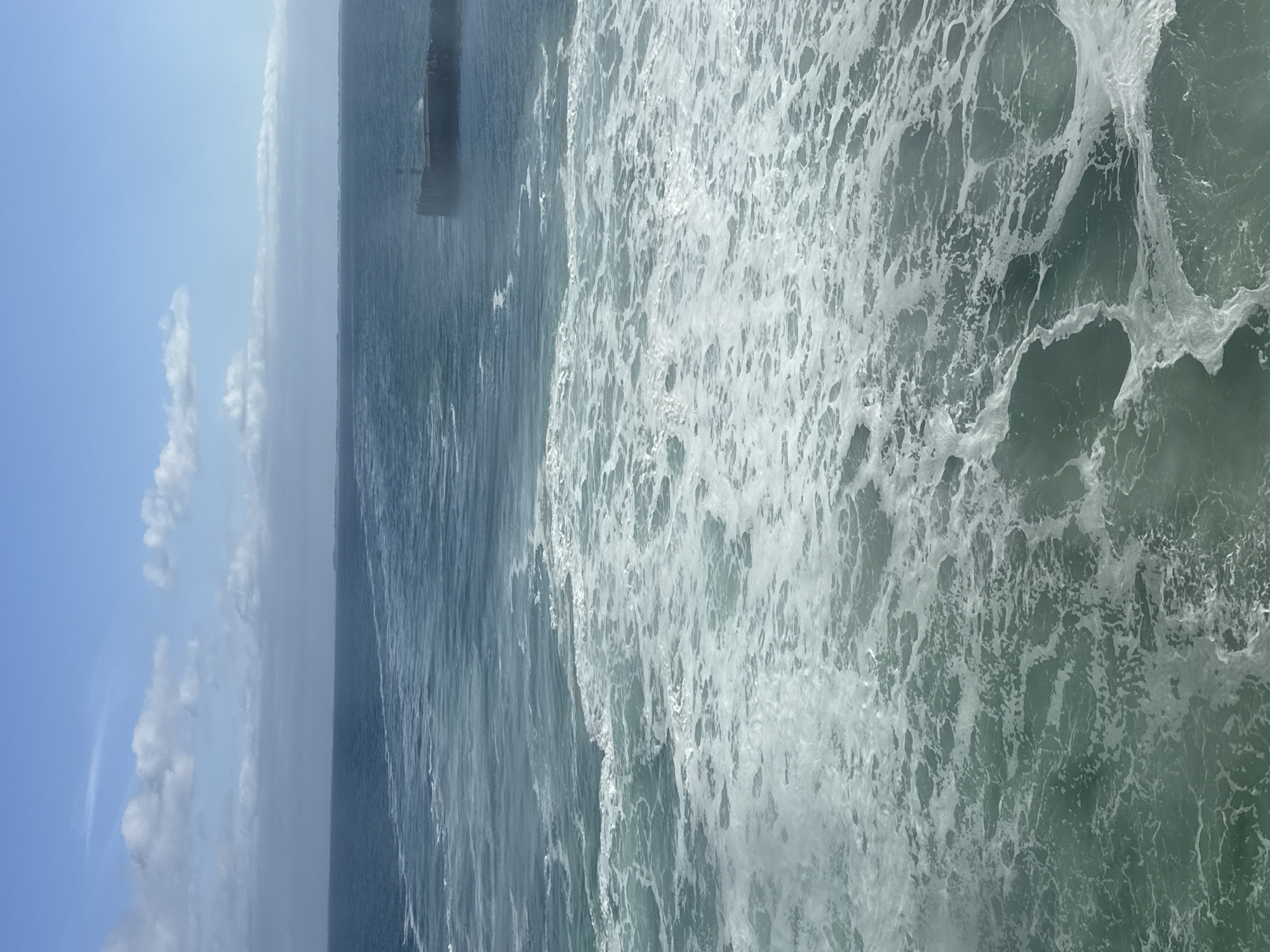 This image captures a choppy sea with white foam churning in the foreground. The water transitions to a darker blue in the distance, meeting a hazy horizon under a bright blue sky with scattered white clouds. In the middle distance, on the right side of the frame, a dark, rectangular structure, possibly a pier or breakwater, juts out into the sea. The scene suggests a possibly windy day at sea, with no people or specific landmarks clearly visible to identify a precise location within Unknown, Denmark.