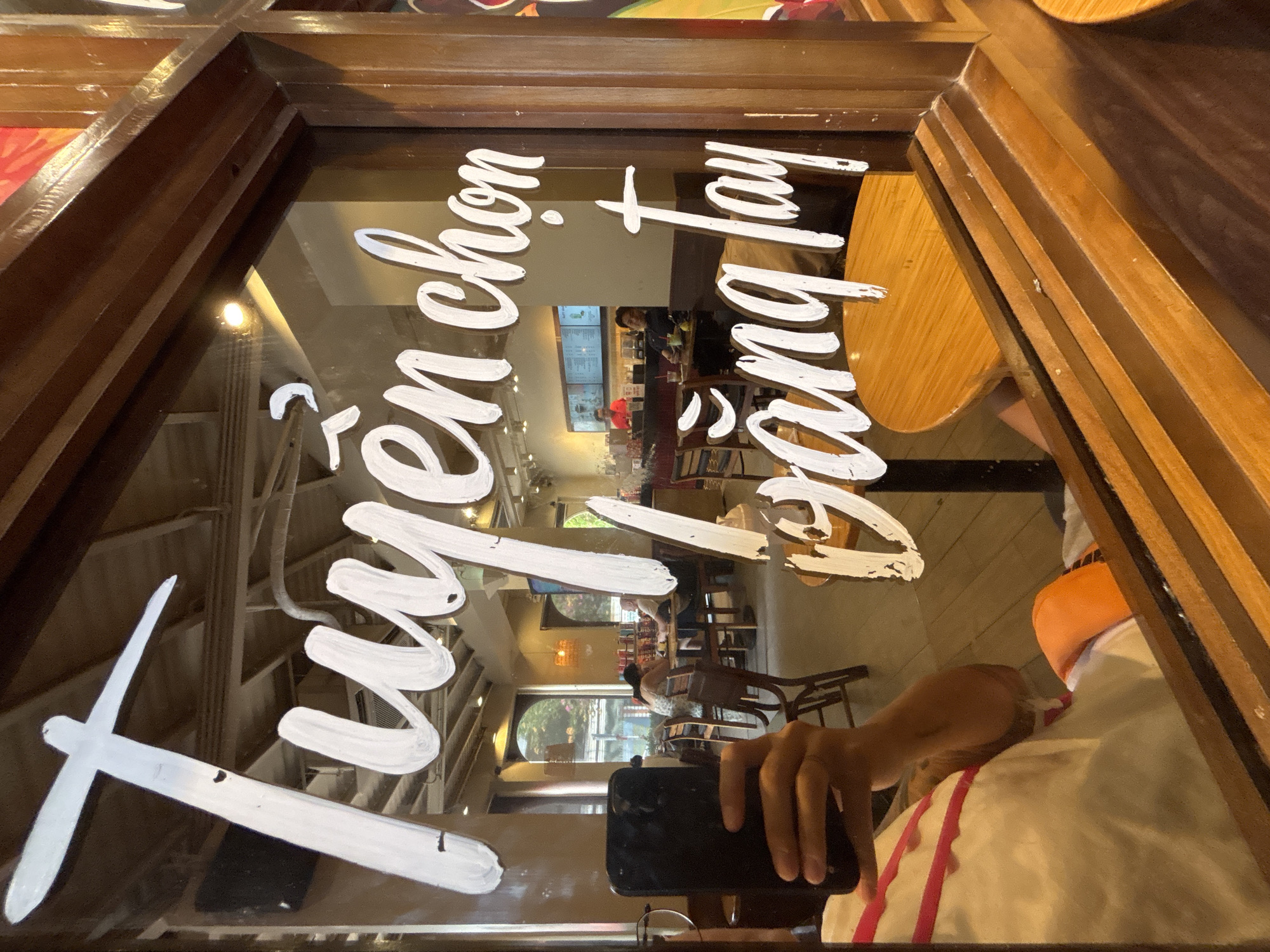 The image, taken at a coffee shop in Nha Trang Ward, Vietnam, shows a reflection on a glass window. The reflection displays white lettering that reads "Tuyển chọn vang tay." The text appears to be handwritten with a brushstroke effect. In the foreground, a person is holding a black smartphone, possibly taking the picture. The background of the reflection reveals the interior of the coffee shop, with patrons sitting at tables and a counter with staff behind it. The overall lighting suggests it is daytime.