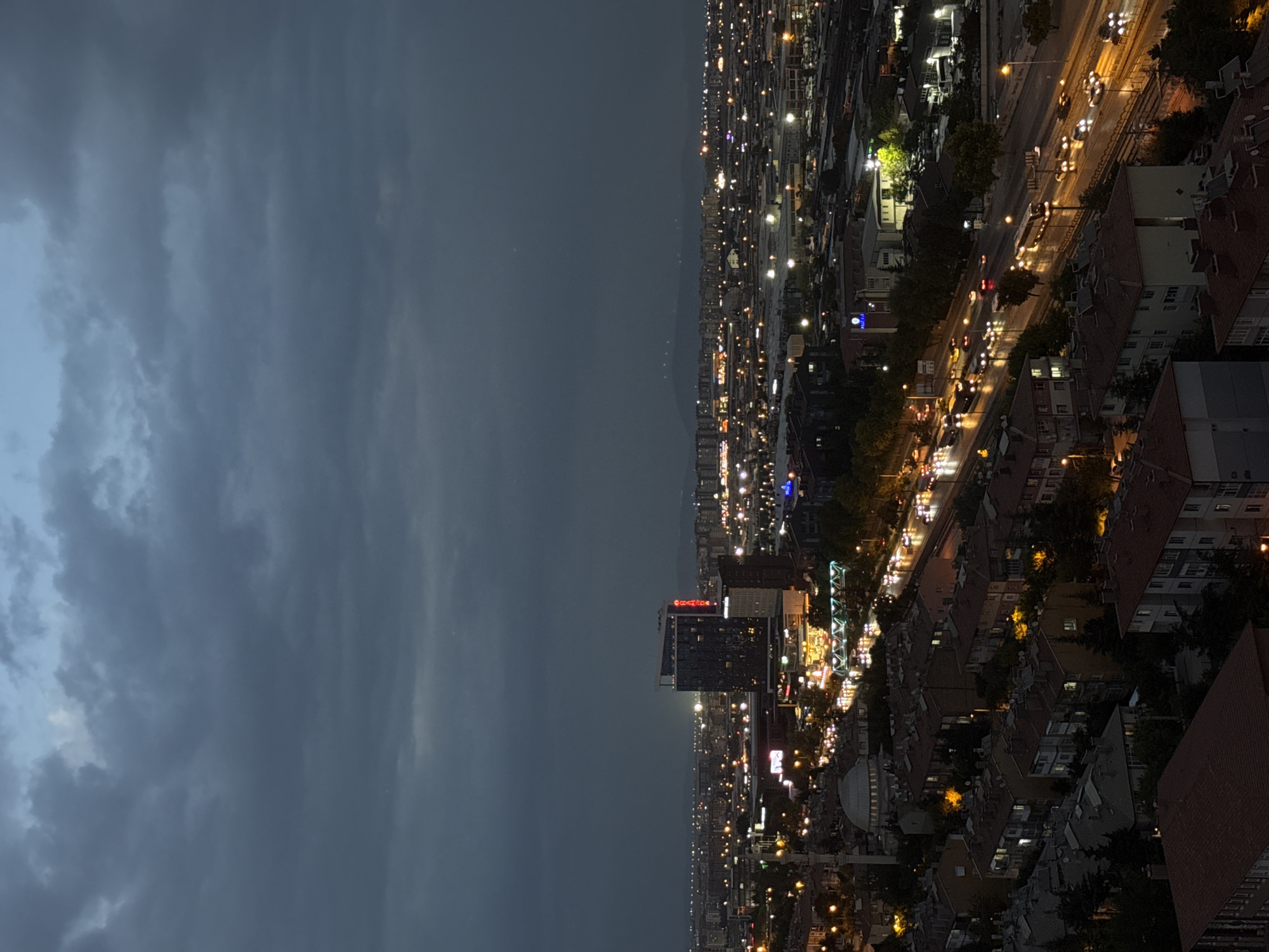An aerial nighttime view captures a section of the city of Konya, Turkey, under a dark, cloudy sky. The scene is dominated by the glow of city lights reflecting off the wet pavement of a multi-lane highway. Numerous vehicles, their headlights and taillights creating streaks of light, are visible on the road, indicating moderate traffic.
In the mid-ground, a prominent modern building with illuminated signage stands out. To its left, a domed structure, likely a mosque, is visible, along with other residential buildings and smaller commercial structures. The foreground consists of densely packed residential houses with red-tiled roofs. Trees and streetlights are interspersed throughout the urban landscape. The overall impression is one of a bustling city at dusk or after dark, with the weather contributing to a dramatic atmosphere.